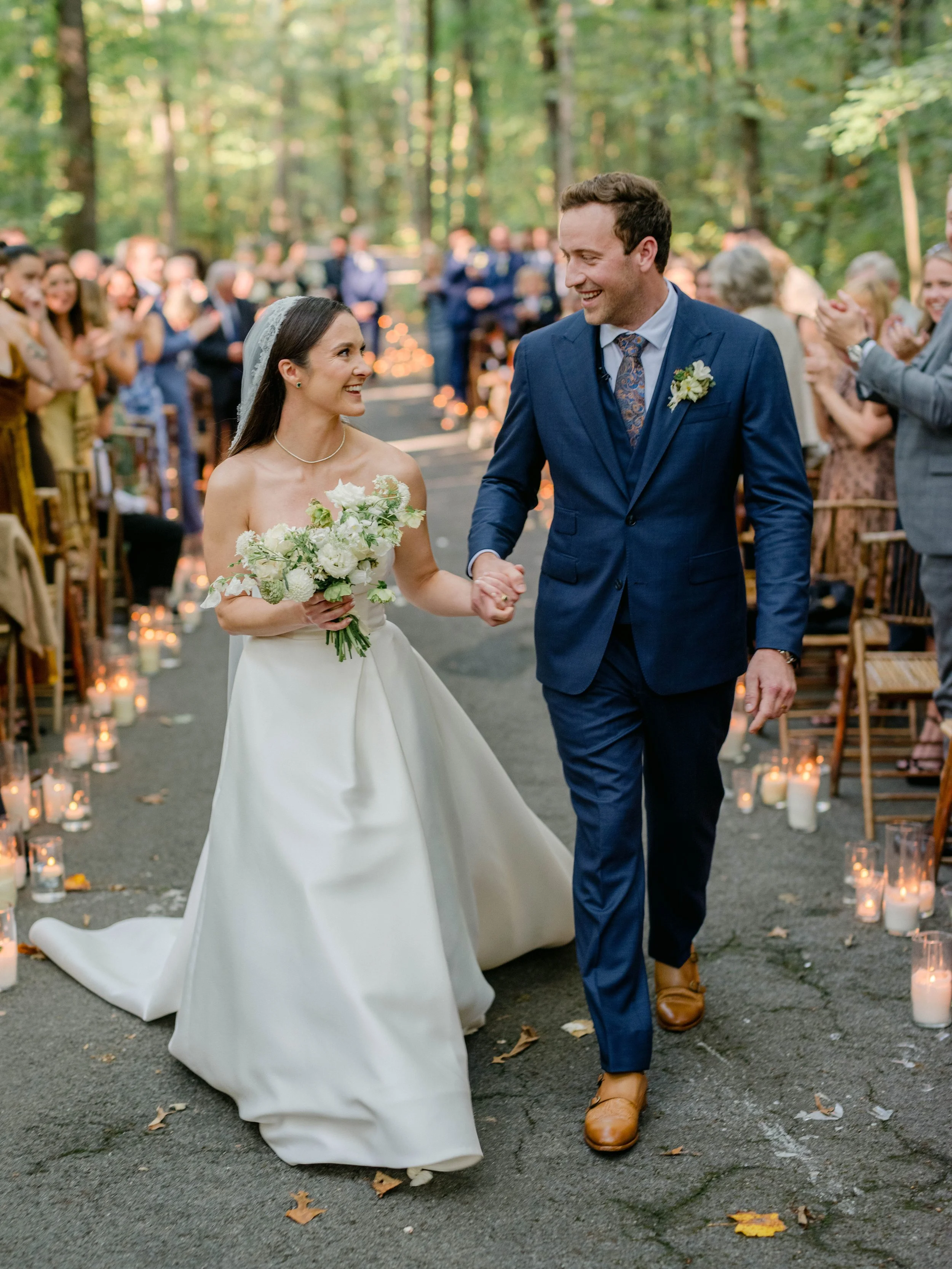 A bride and groom walking hand in hand down the outdoor aisle, smiling at each other, surrounded by wedding guests, in a forest setting with candles lining the path.