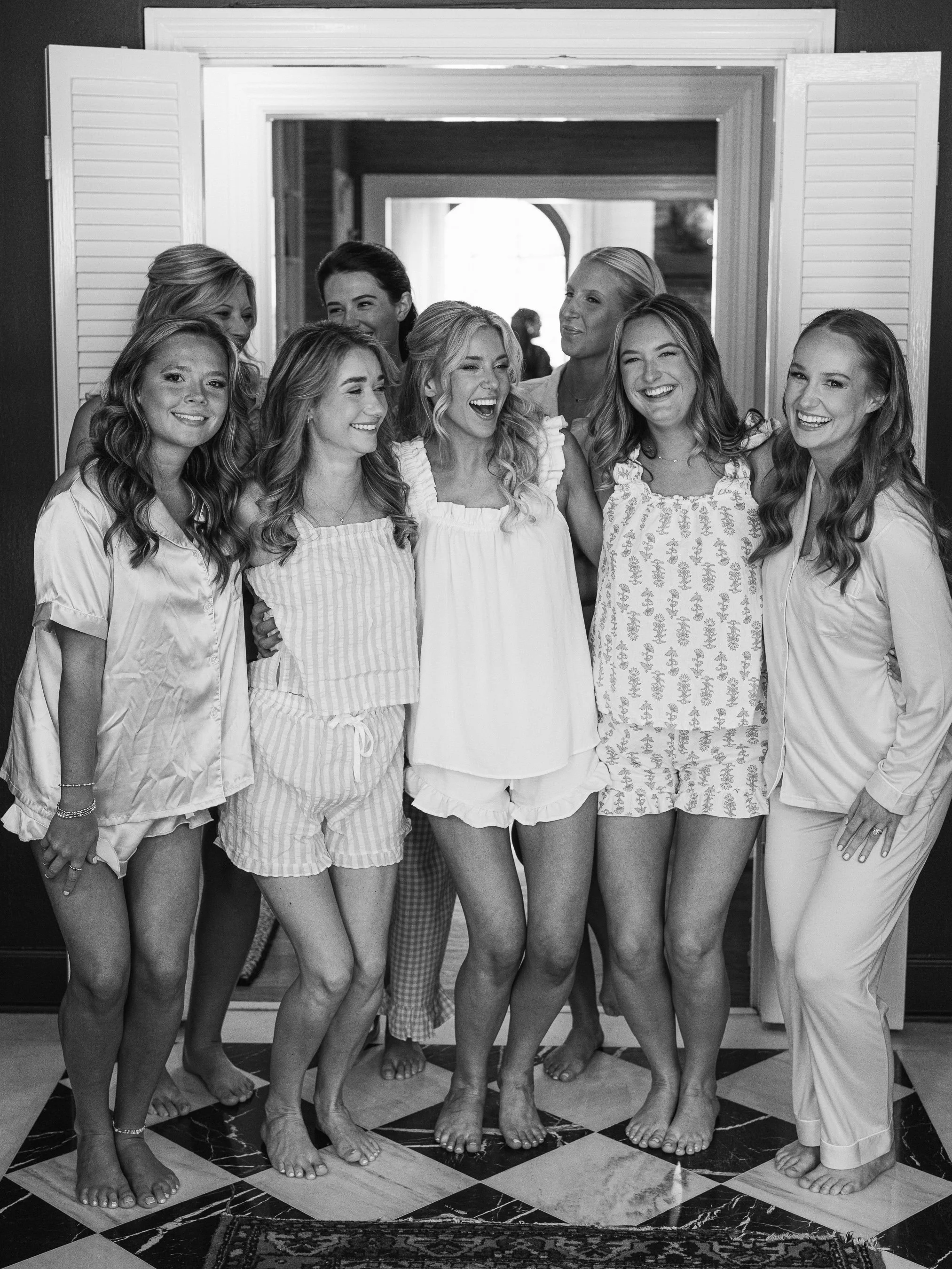 A group of women in pajamas standing together, smiling and laughing, in a cozy indoor setting.