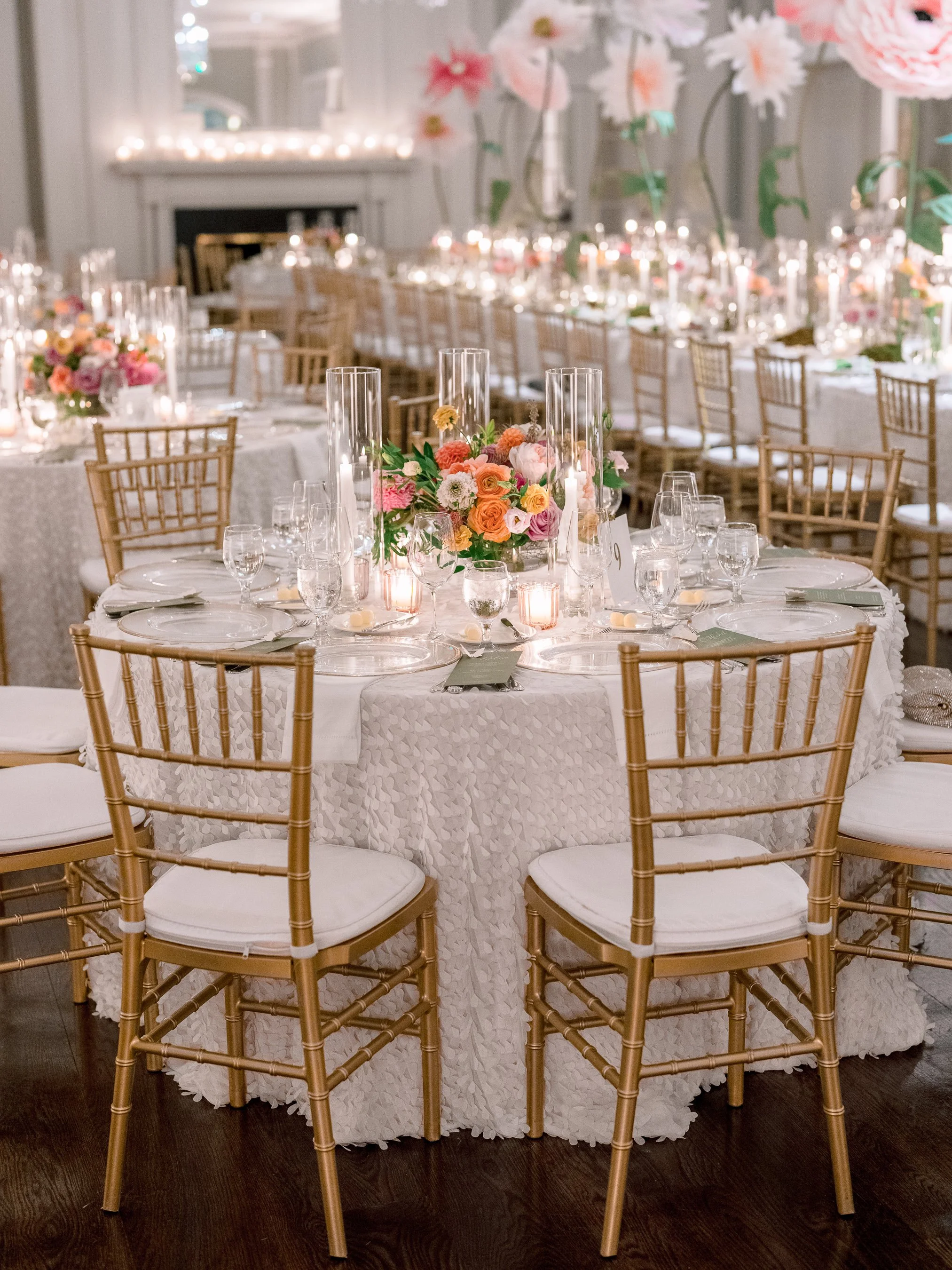 Elegant banquet table decorated with pink, peach, and white flowers, surrounded by gold chairs with white cushions, set with wine glasses, candles, and tableware in a luxurious, softly lit room with floral arrangements.