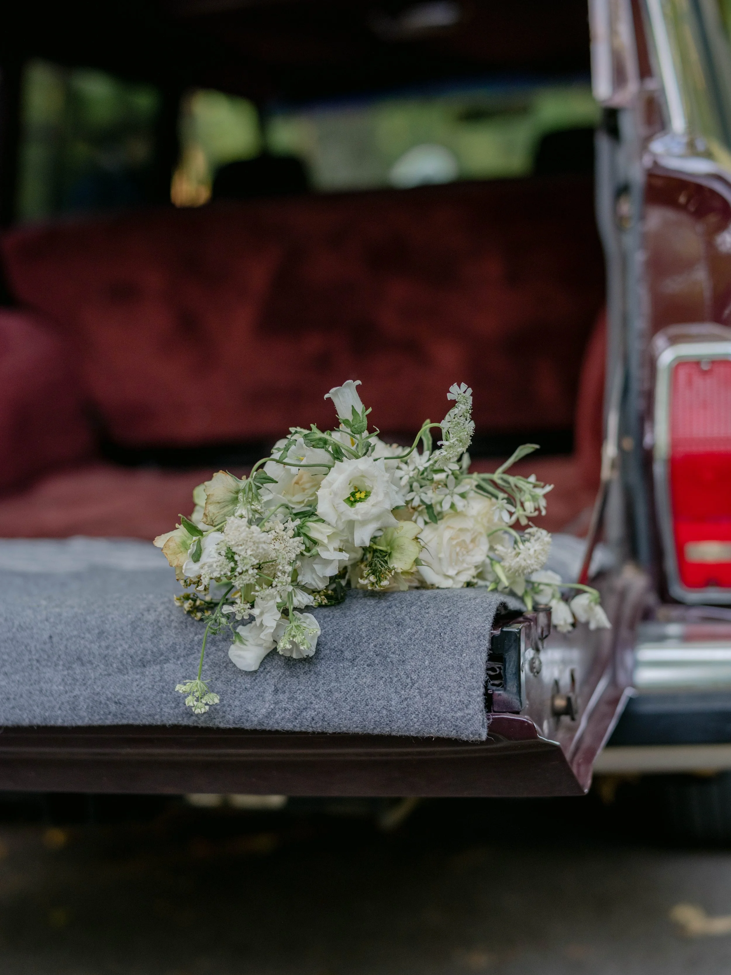 A bouquet of white flowers resting on a gray felt cloth in the open trunk of a vintage dark red car.