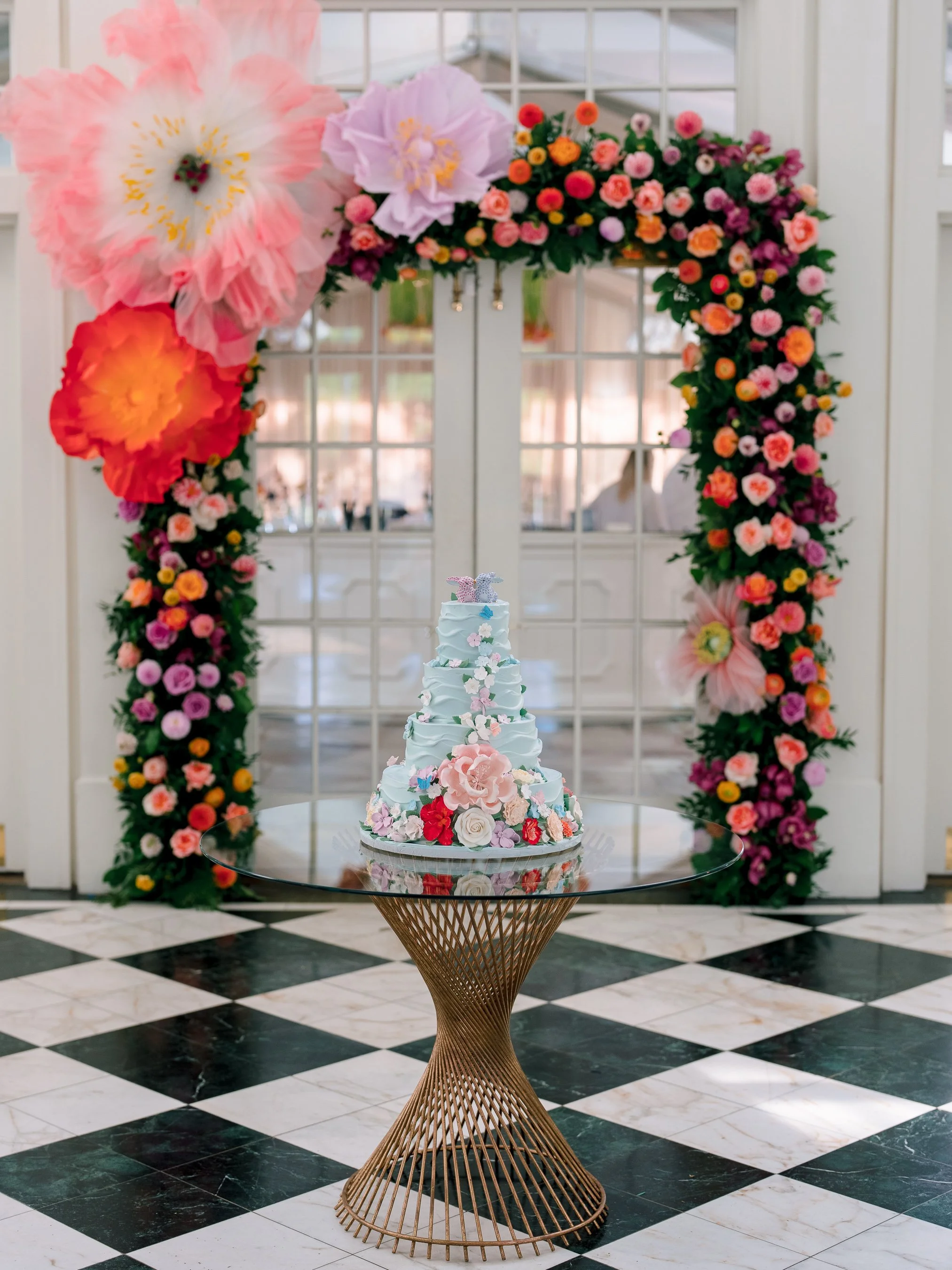 Elegant wedding cake decorated with pink and white flowers on a glass table, with a colorful floral arch in the background inside an elegant venue.