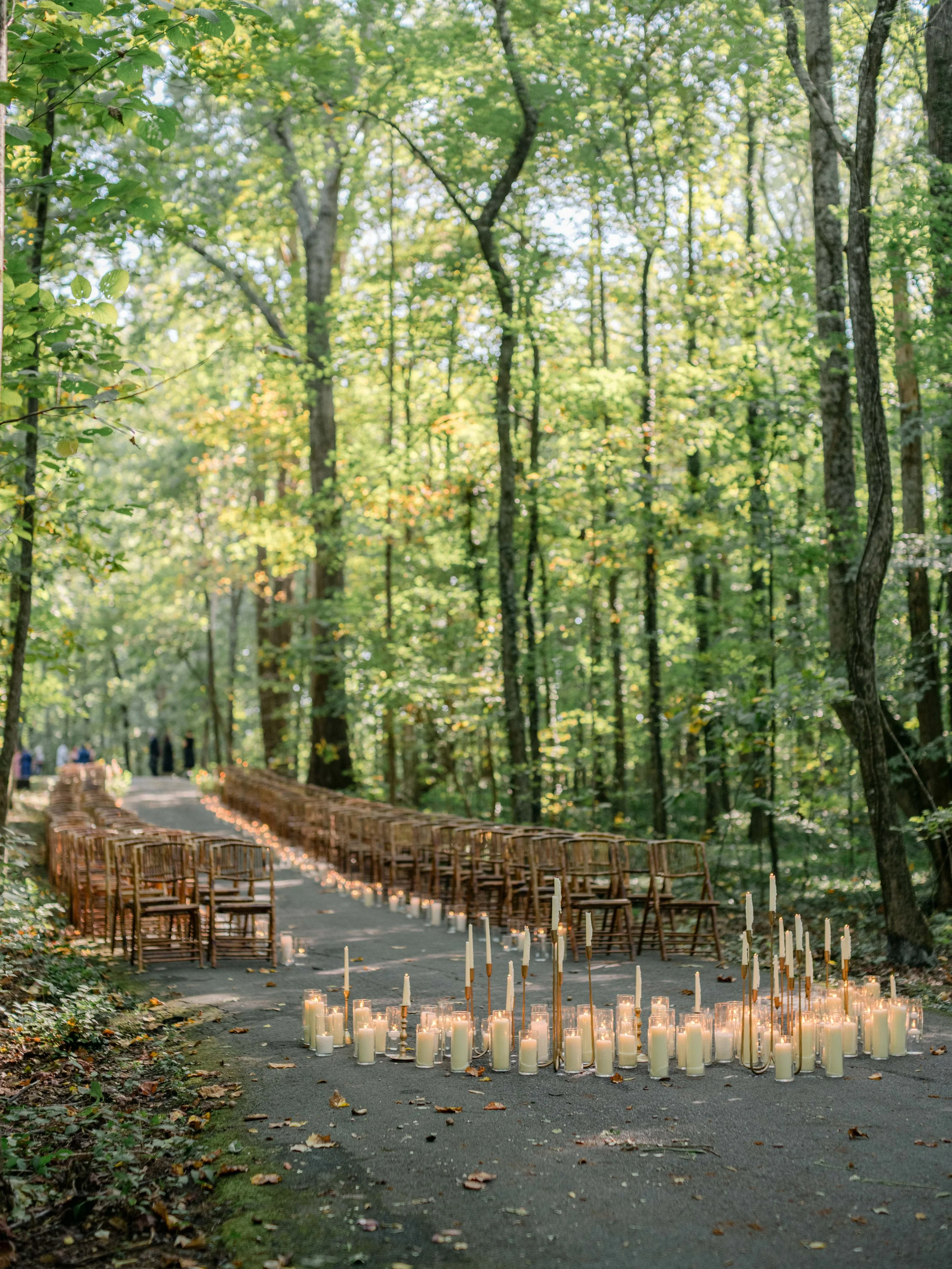 Outdoor wedding ceremony setup in a forest with chairs arranged along a ceremony path, decorated with lit candles and tapers.