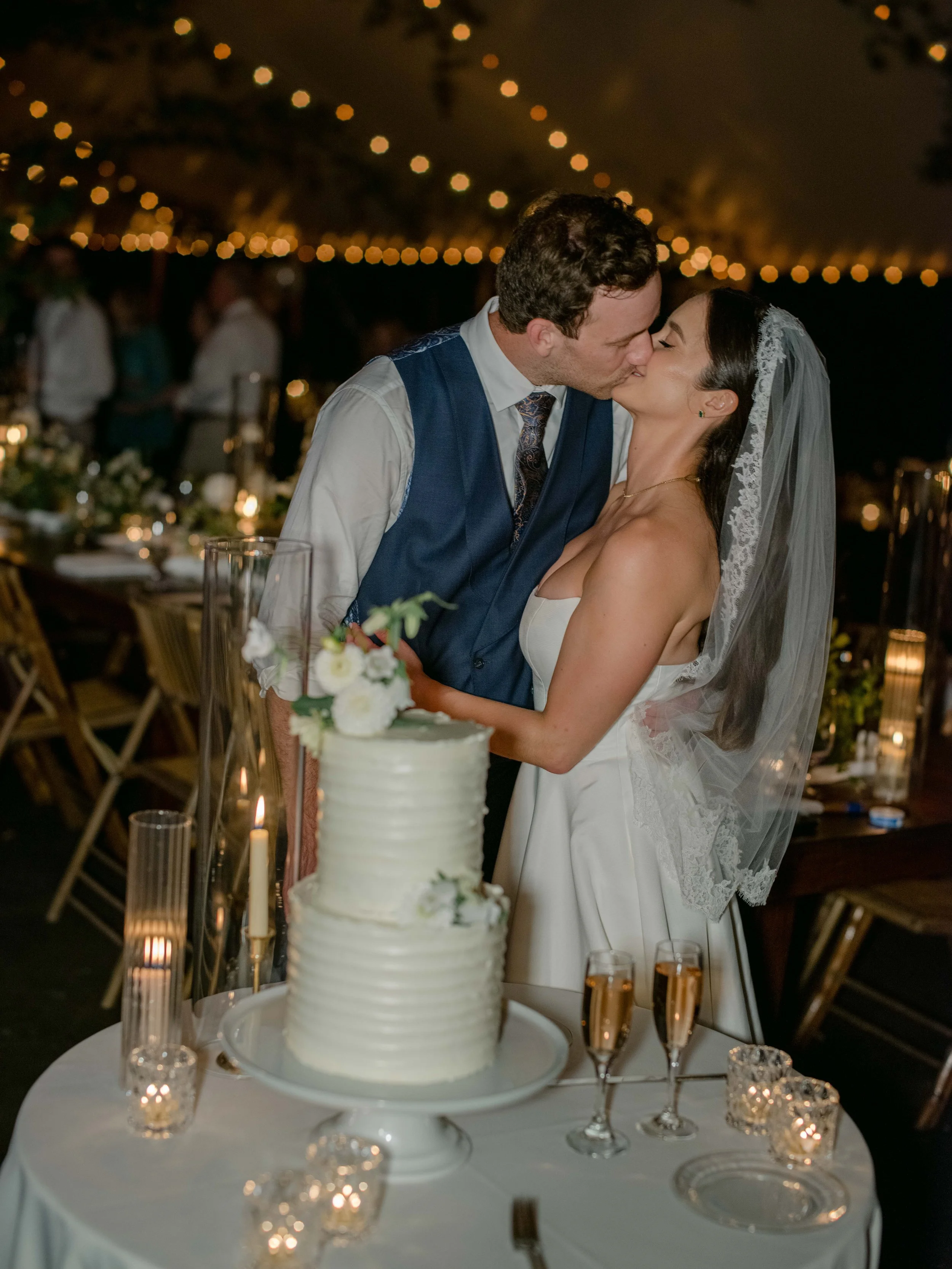 A bride and groom kiss at their wedding reception under string lights, standing behind a wedding cake with candles and champagne glasses on a table.