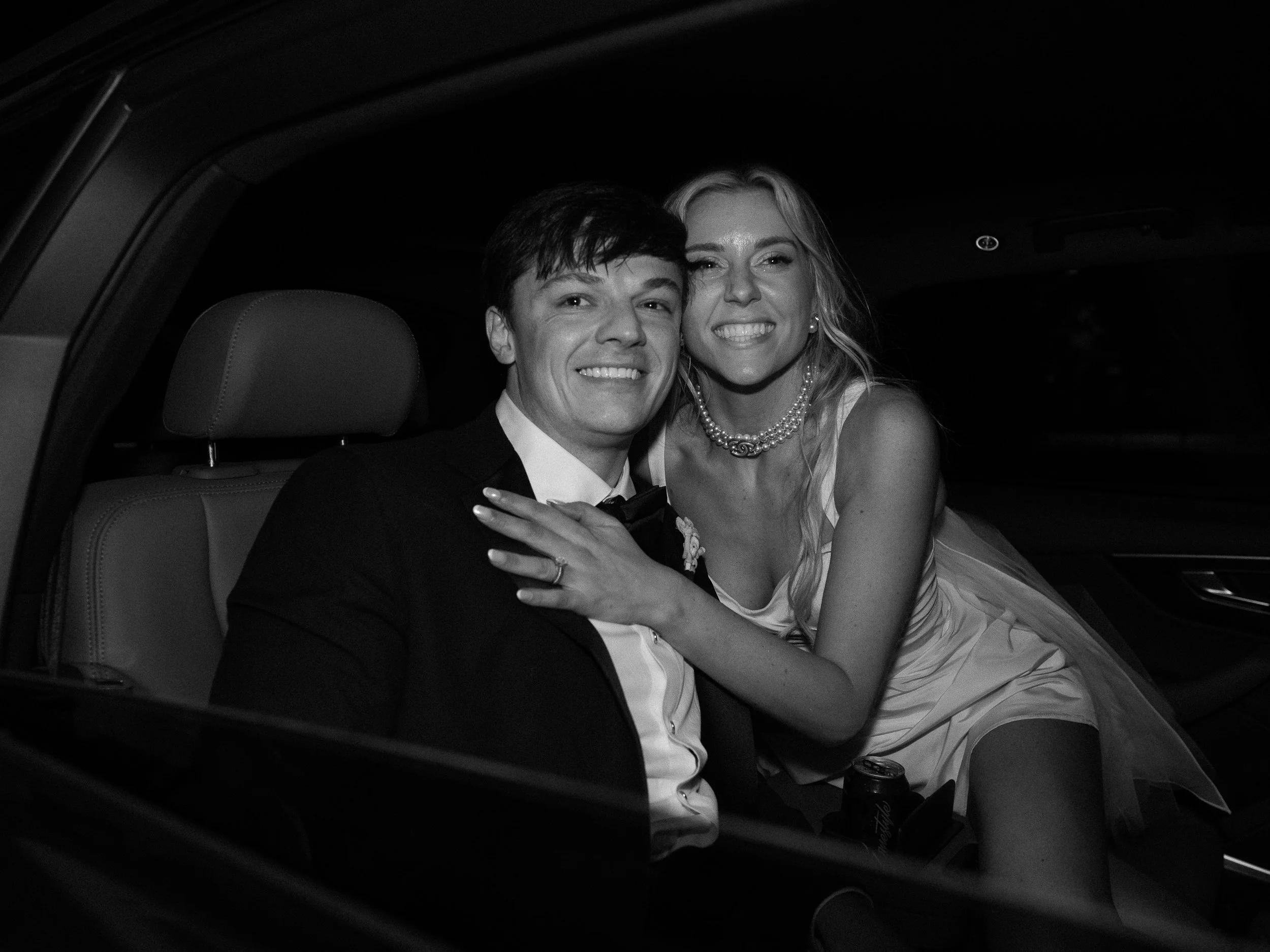 Black and white photo of a joyful couple in wedding attire sitting in a car, smiling at the camera.