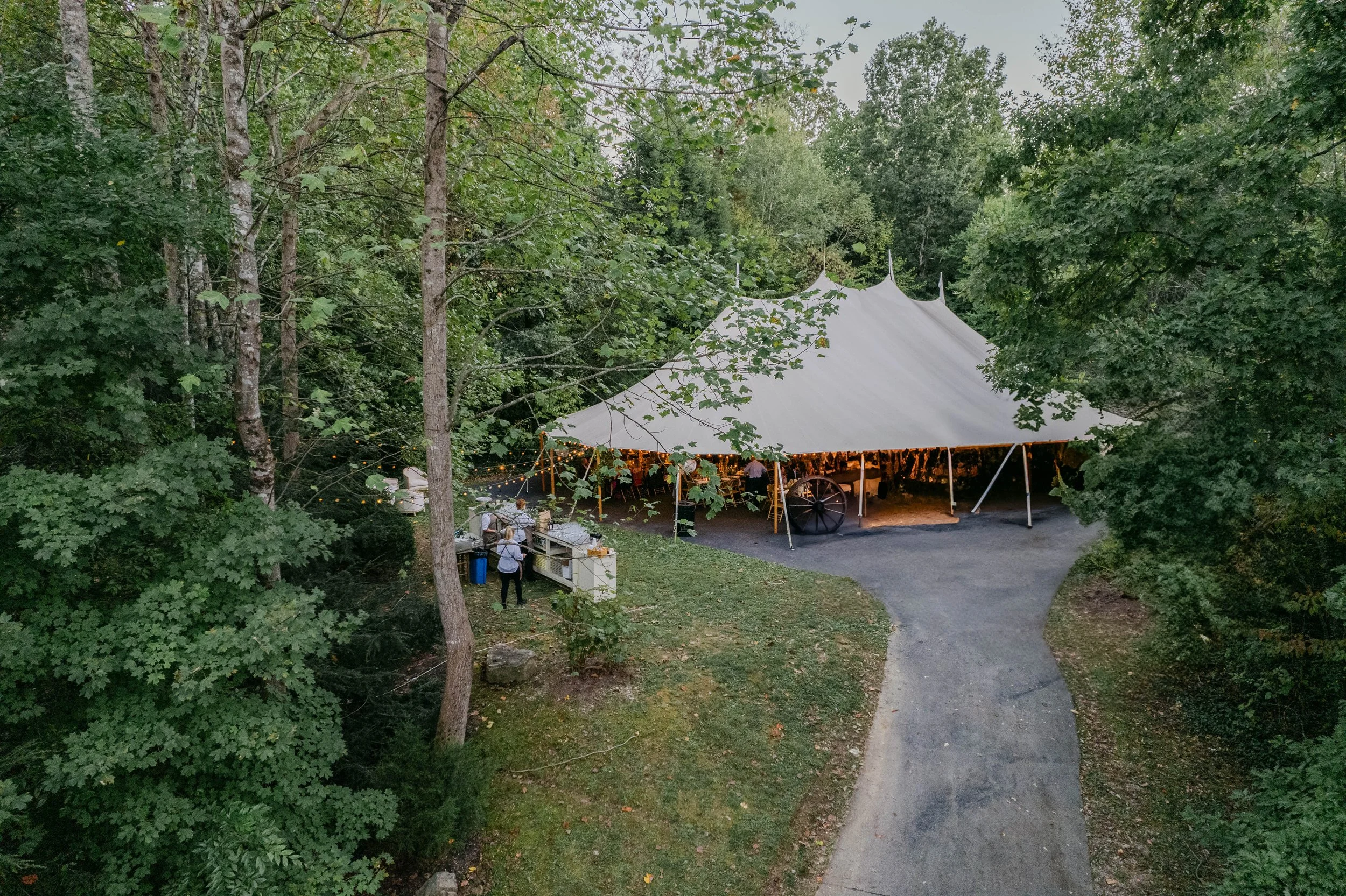 A large white event tent set up in a wooded outdoor area with a gravel pathway leading to it. There are string lights and people near a nearby refreshment or food stand.