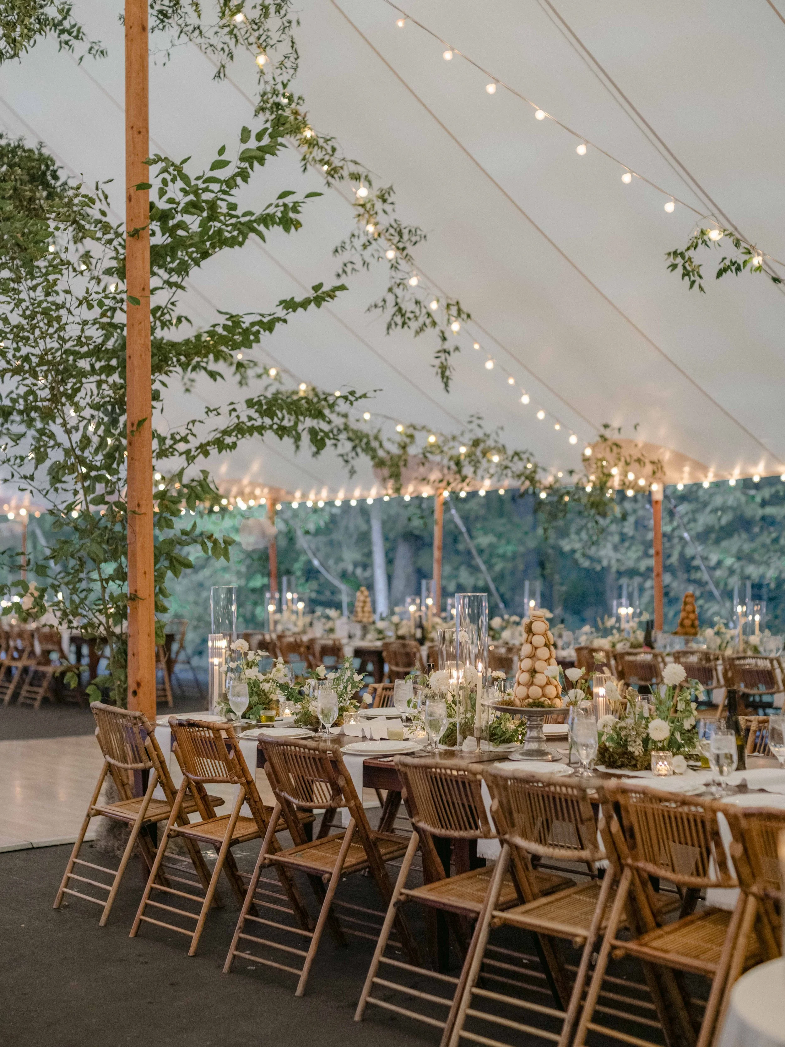 An outdoor event under a tent with string lights, wooden chairs, and rectangular tables decorated with flowers, candles, and macaron towers.