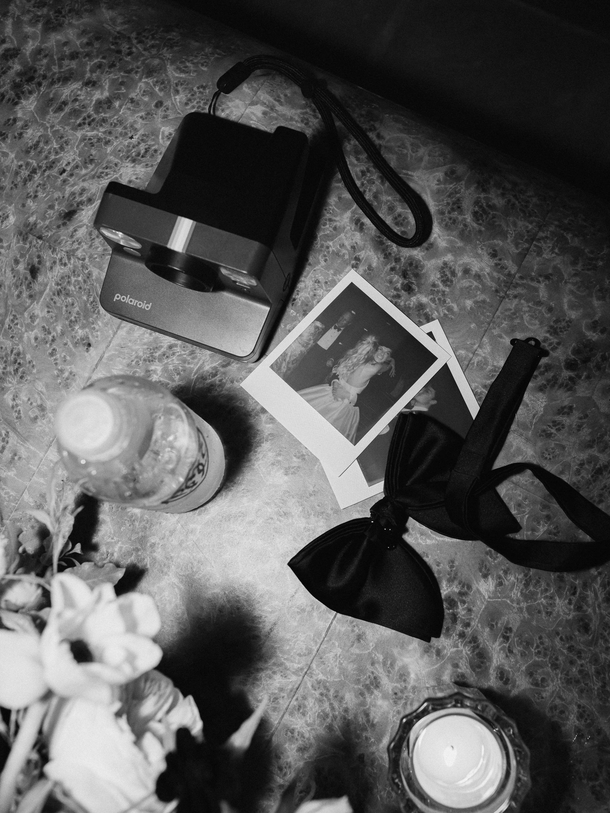 Black and white photograph of a table with a Polaroid camera, two instant photos, a water bottle, a black bow tie, a lit candle, and a floral arrangement.