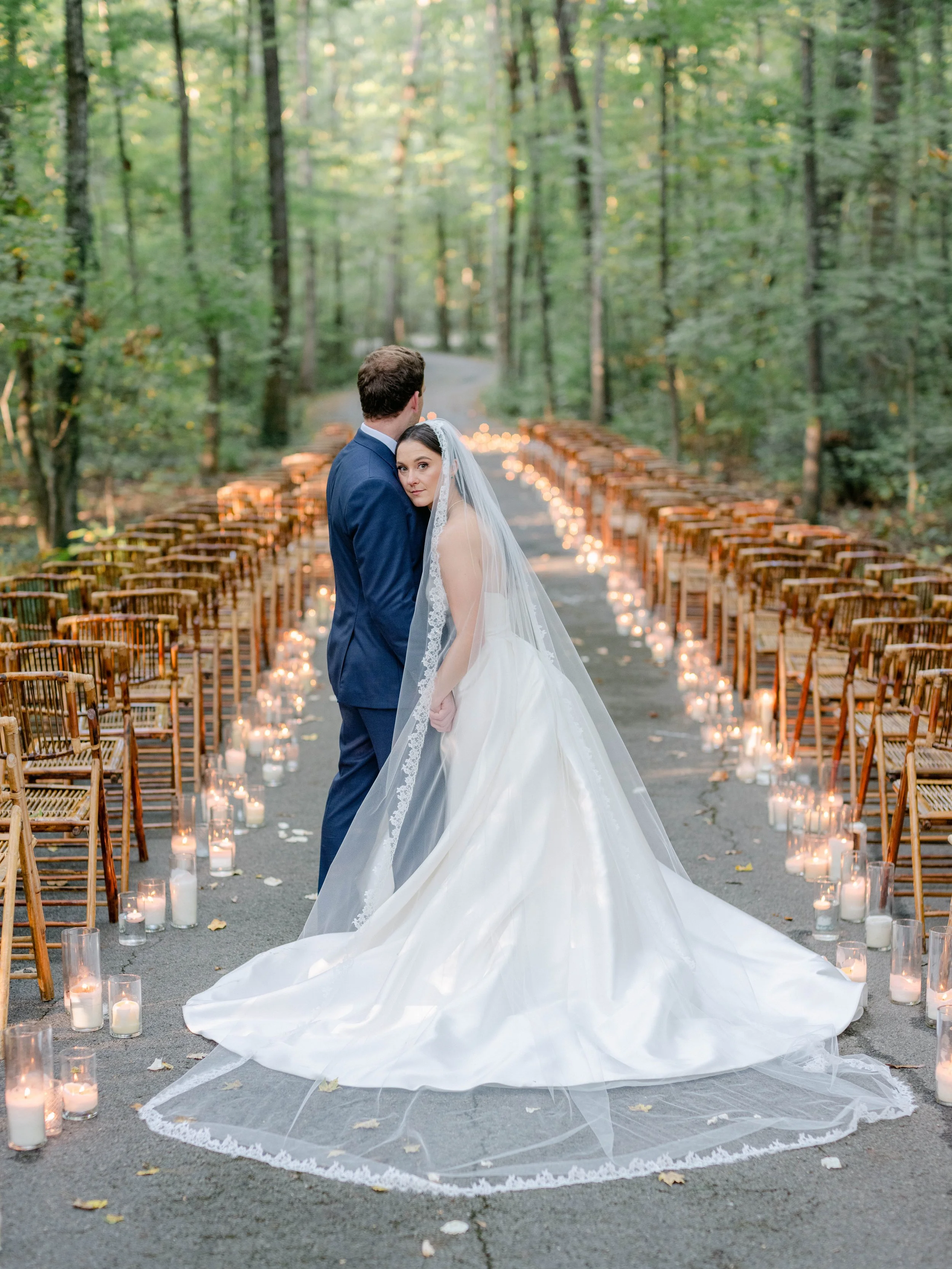 Bride and groom standing closely together in a forested outdoor wedding venue, surrounded by chairs and candles.