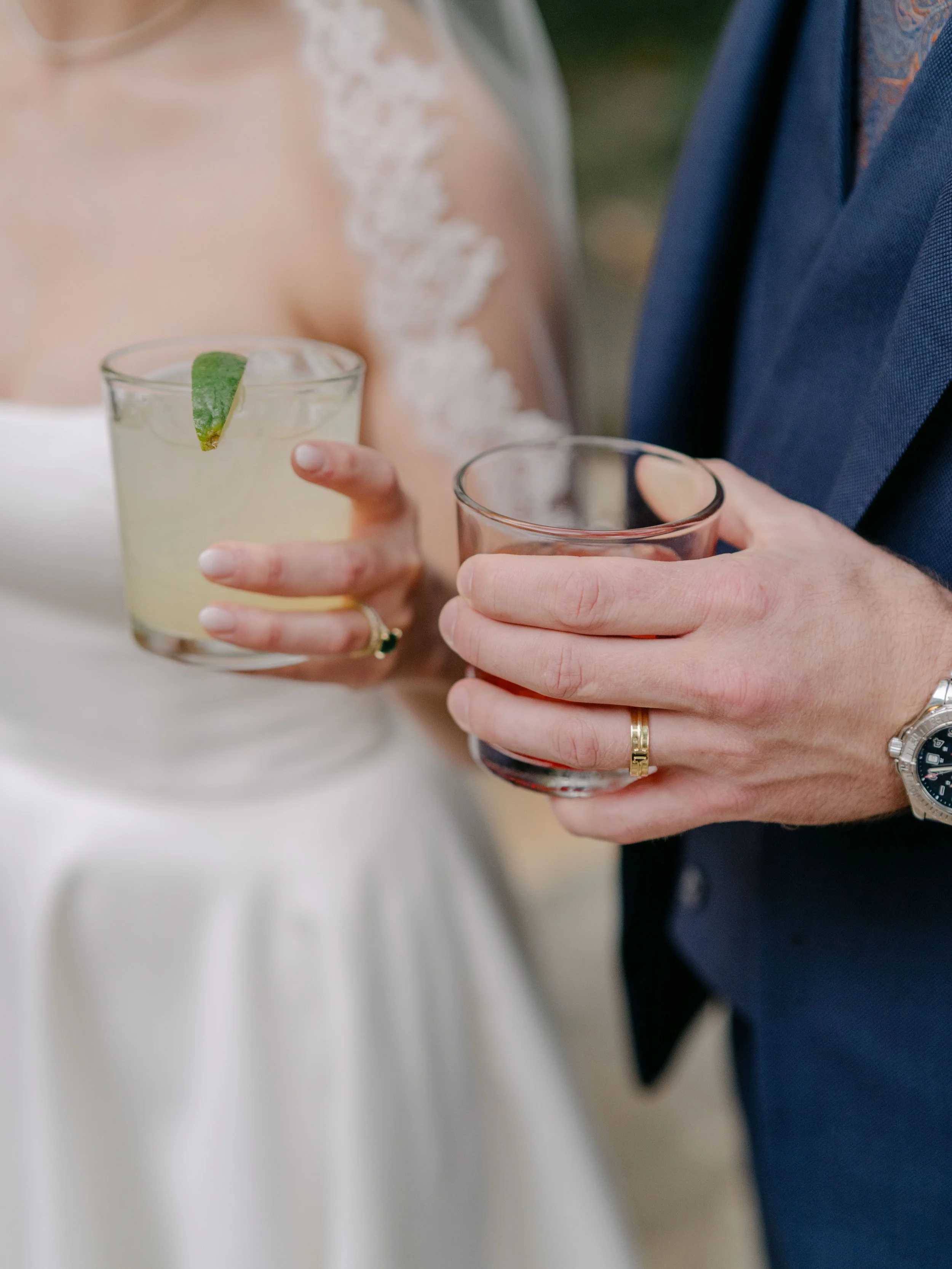 Close-up of a bride and groom holding drinks at a wedding reception, with the bride wearing a lace wedding dress and the groom in a dark suit.