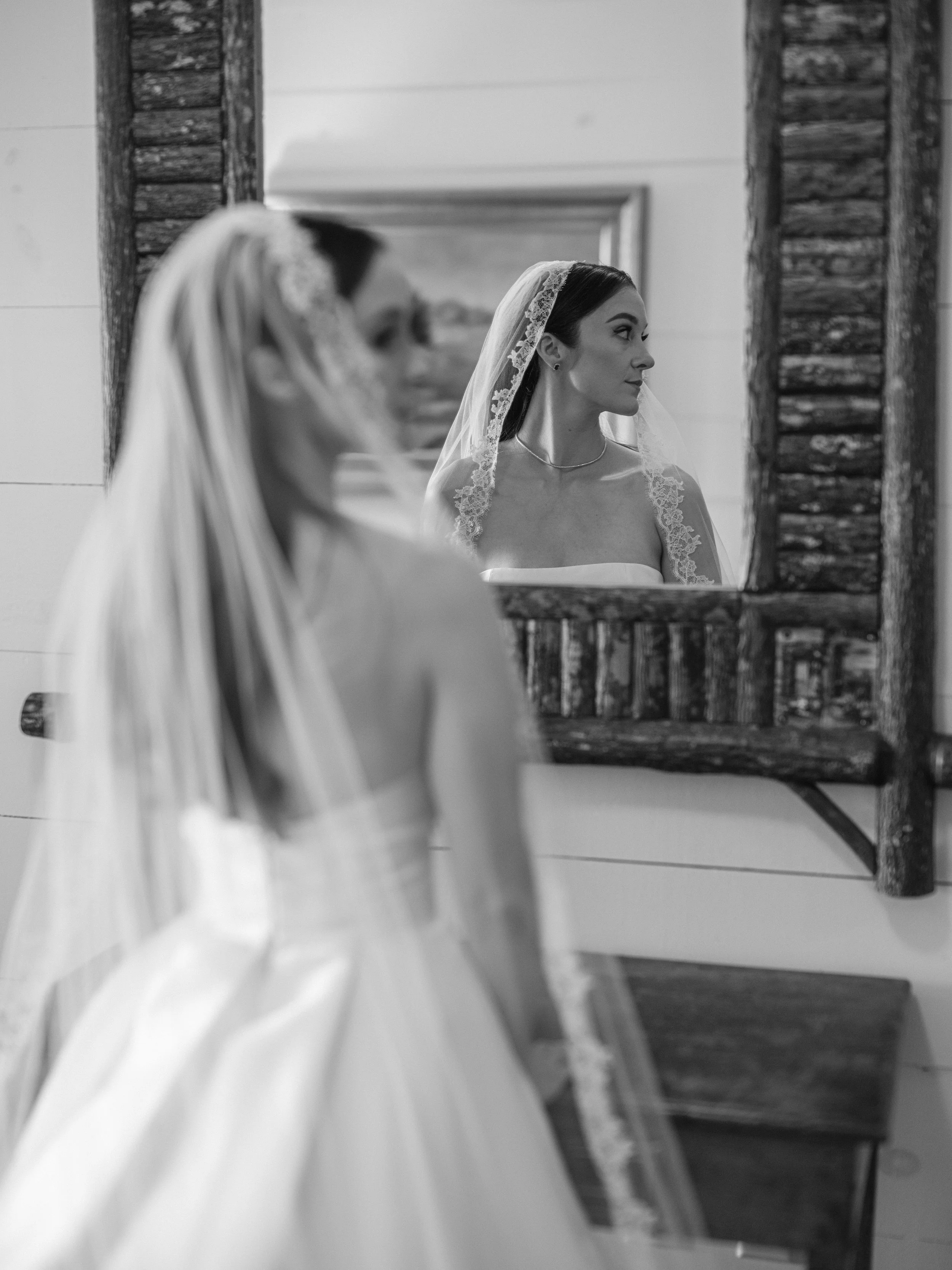 A bride stands in front of a mirror, wearing a wedding gown and veil, with her reflection showing her face and elegant makeup.