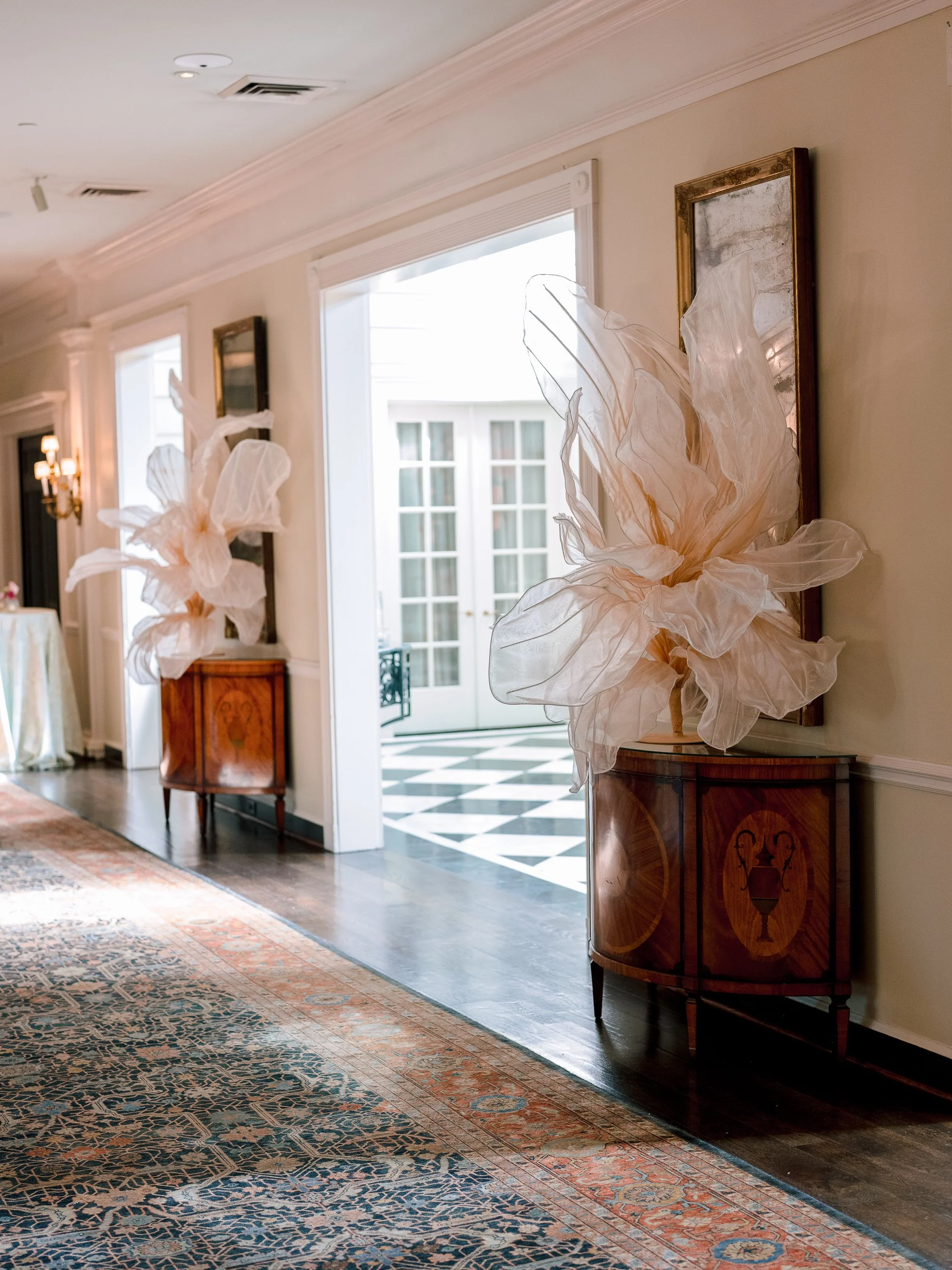 Elegant hallway with two wooden cabinets, each topped with a large, decorative, cream-colored fabric flower sculpture, and framed artwork on the walls.