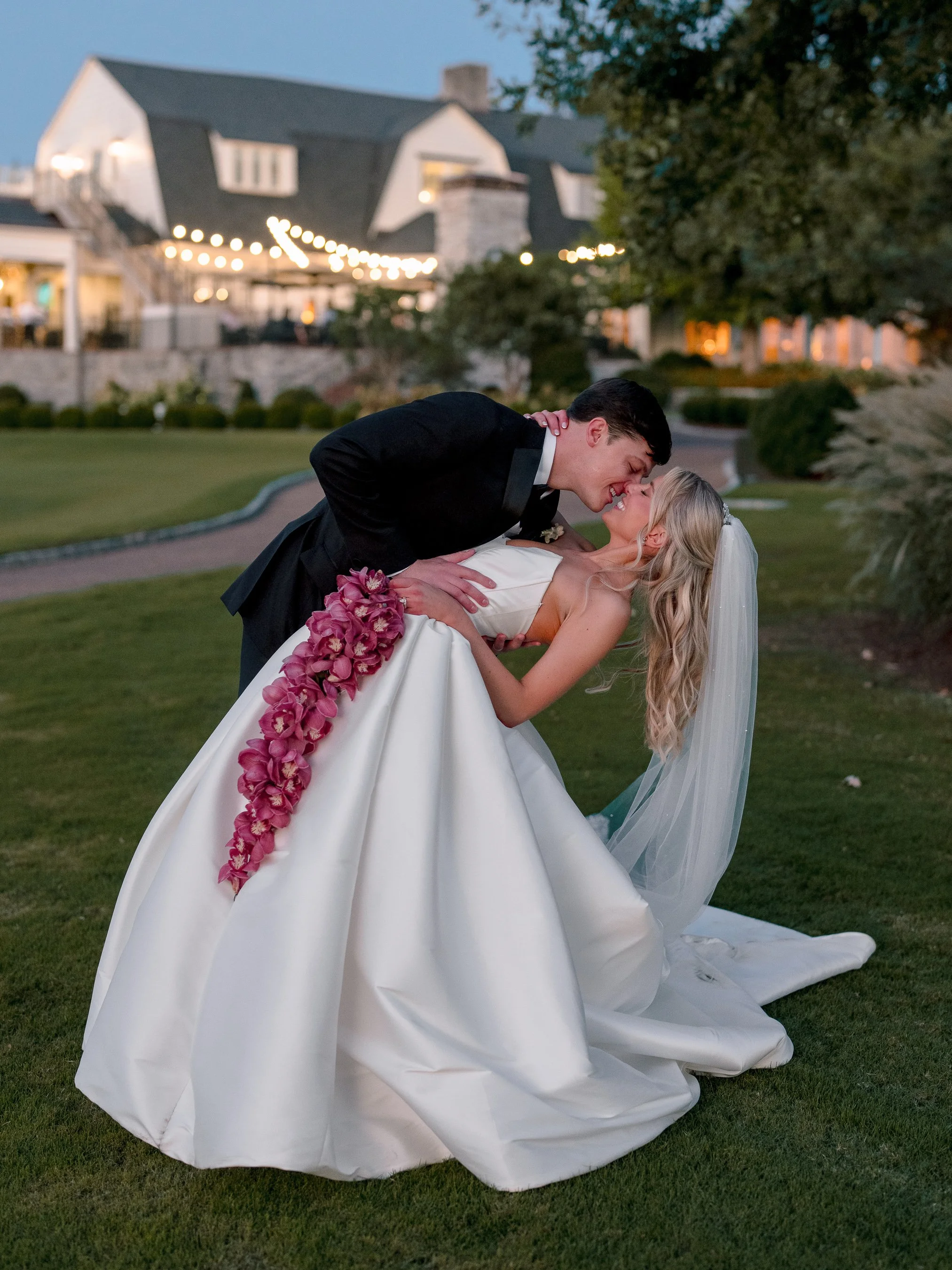 A newlywed couple sharing a kiss outside on a grassy lawn during dusk. The groom is in a black tuxedo, and the bride in a white wedding gown with a long train and veil. The bride's gown has a trail of purple flowers. The background features a large w