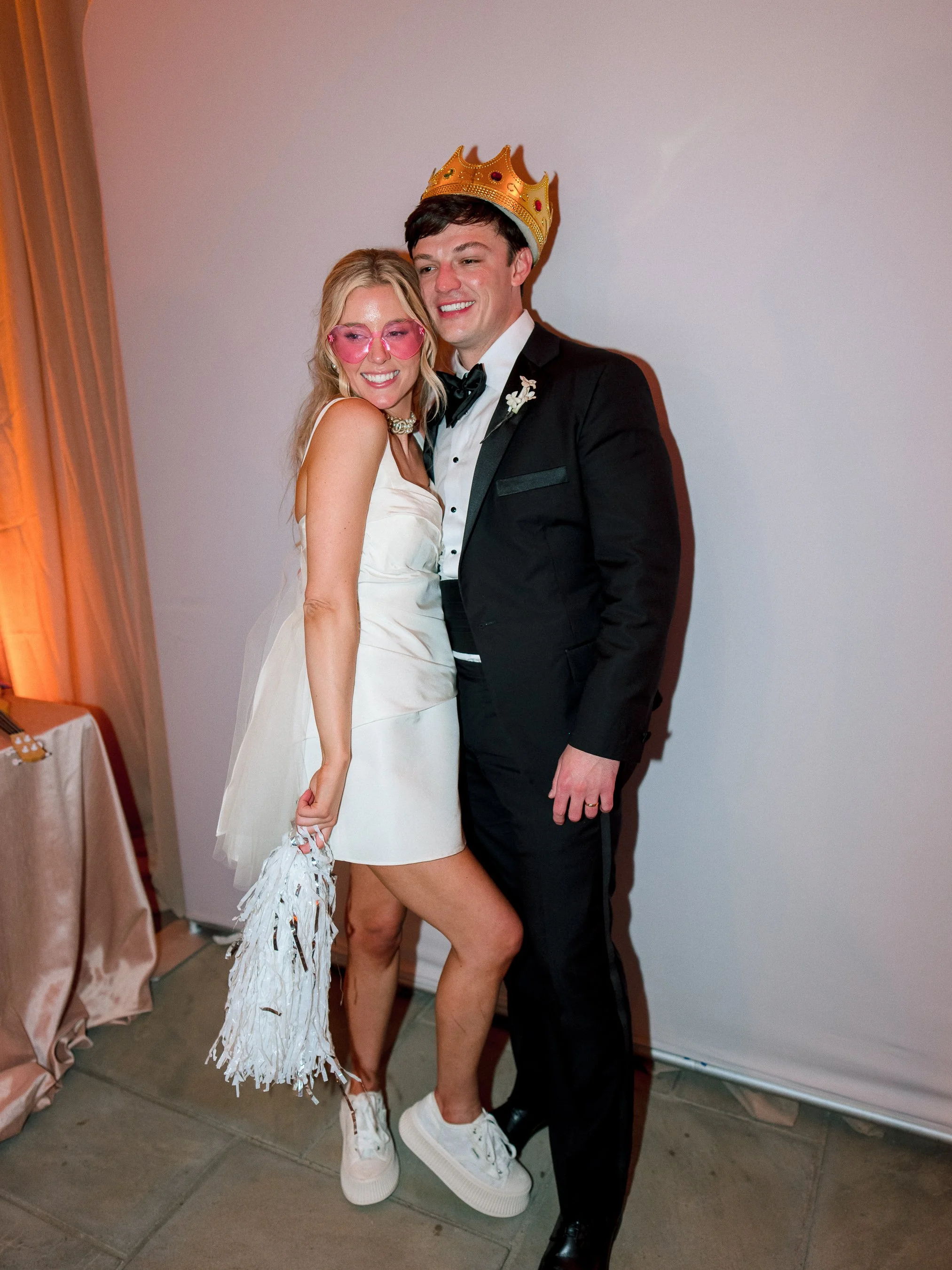 A couple at a celebration, dressed elegantly; the woman wears a white dress, pink sunglasses, and white sneakers, holding a white tassel bag, while the man wears a black tuxedo with a bow tie, and a gold paper crown.