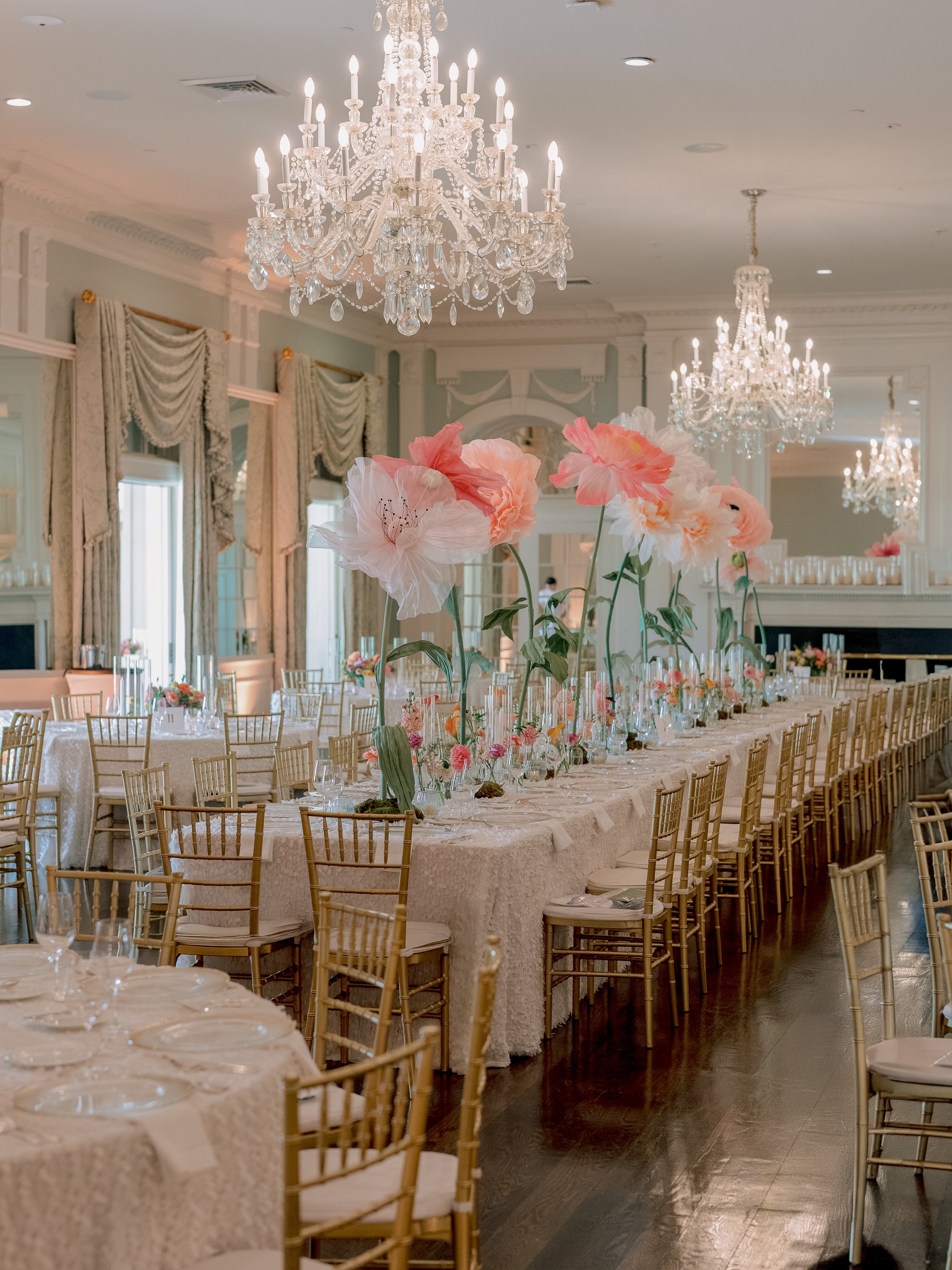 An elegantly decorated banquet hall with ornate chandeliers, tall pink and white floral centerpieces on long tables, and gold chairs arranged for a formal event.