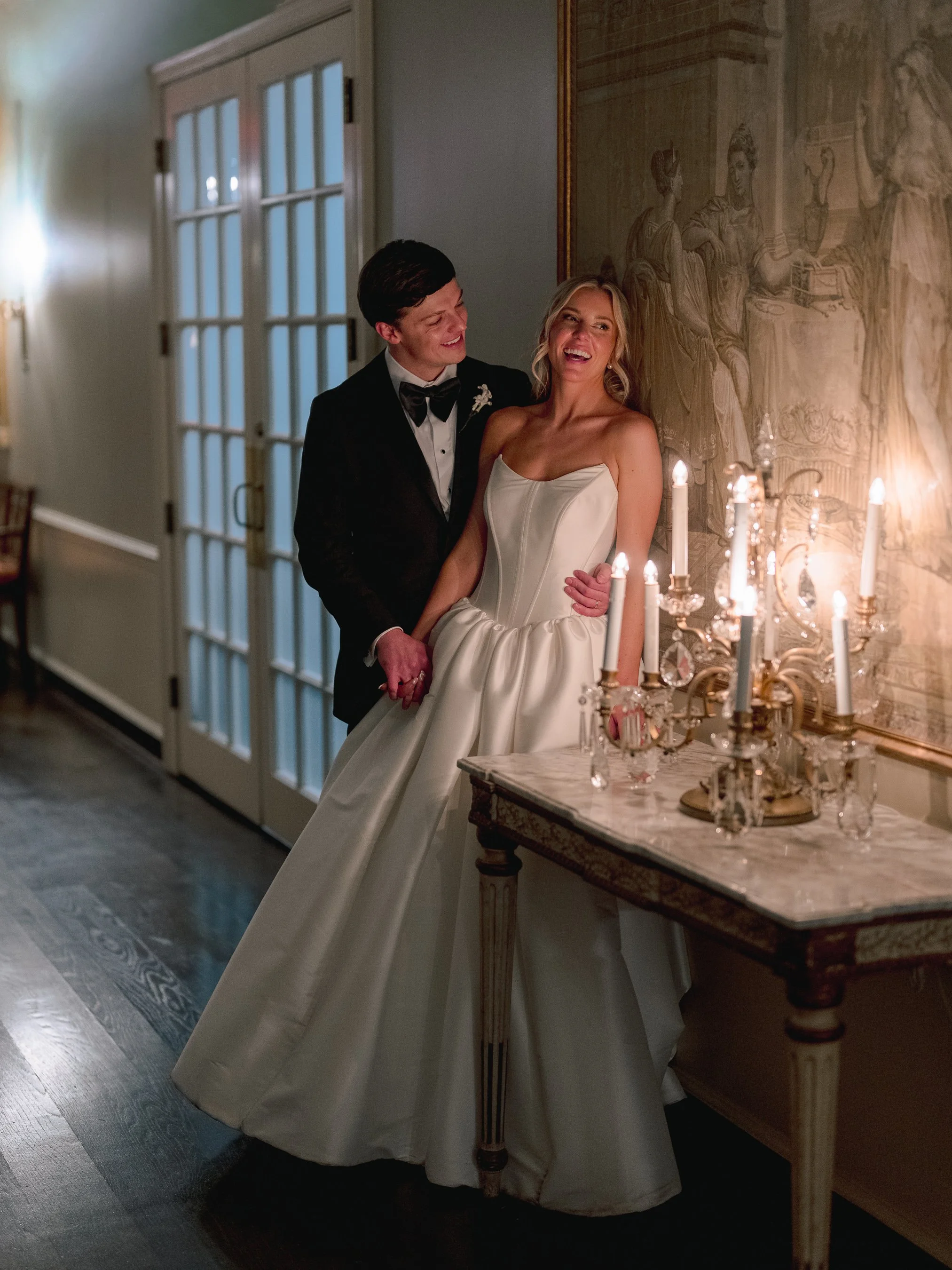 A bride and groom standing together at their wedding reception or ceremony, smiling and looking happy. The bride is wearing a strapless white wedding gown and the groom is wearing a black tuxedo with a bow tie. They are positioned next to a vintage-s
