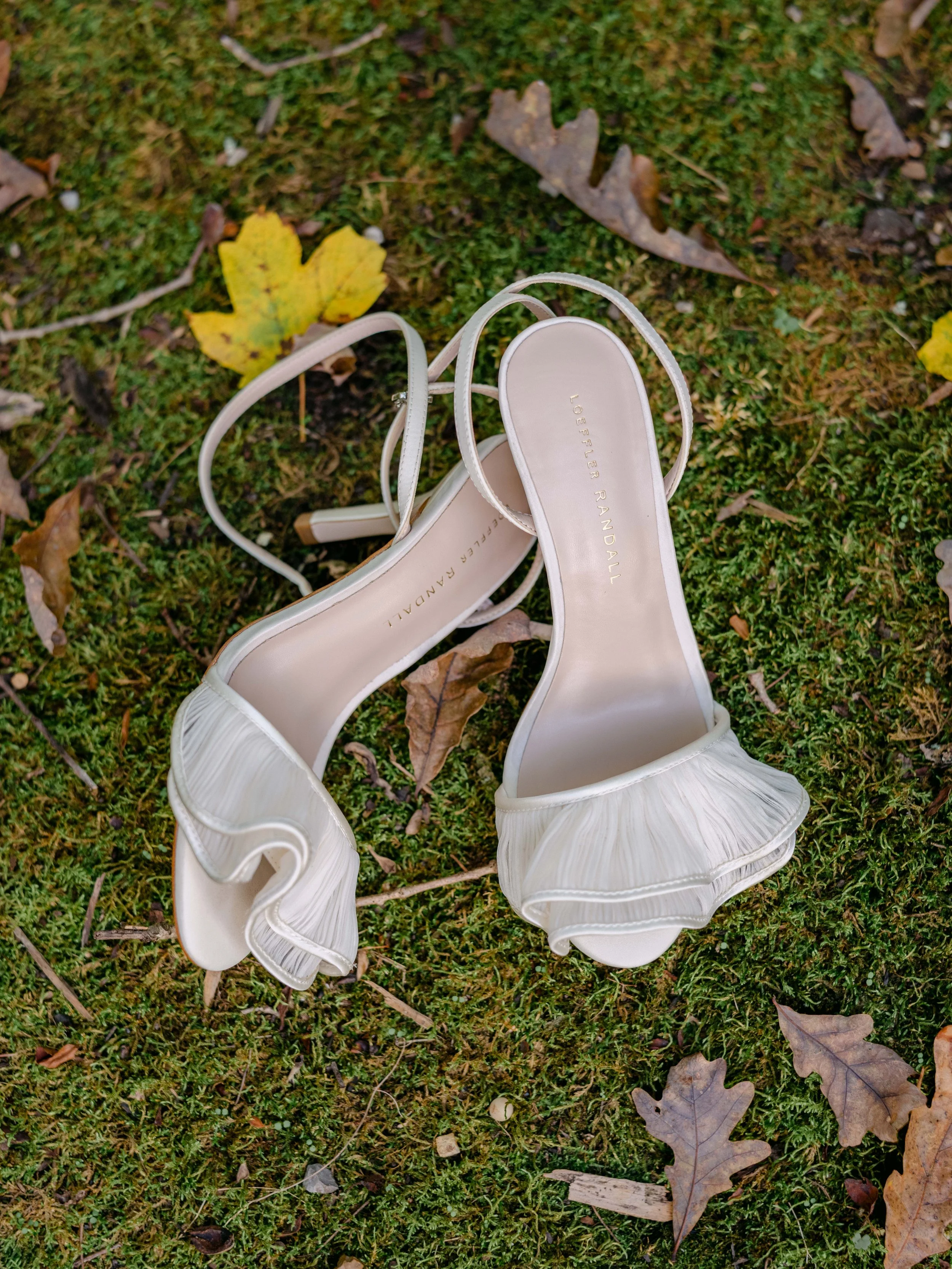 White high-heeled sandals with ruffle details on the straps, lying on a mossy ground surrounded by fallen leaves.