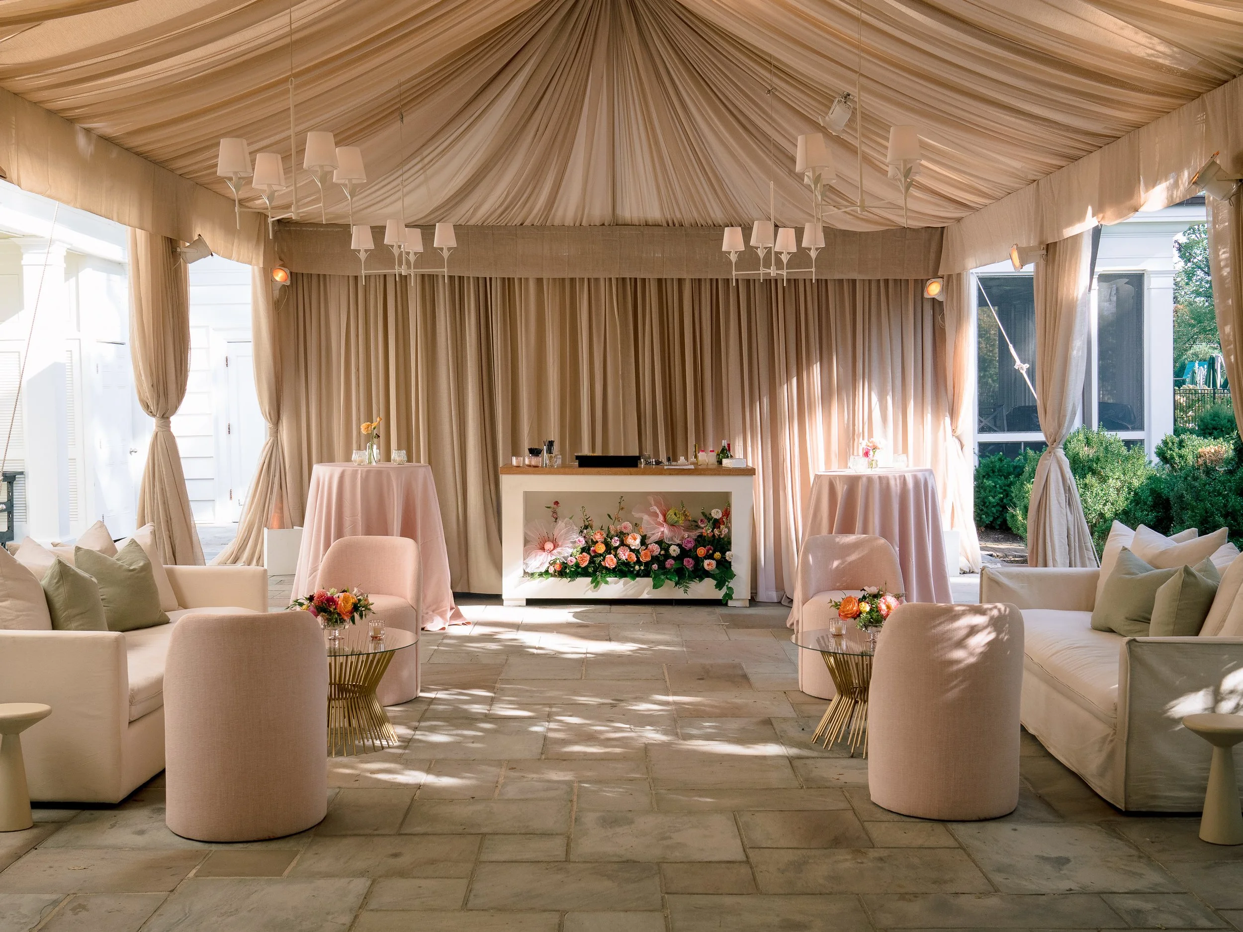 Elegant reception area with beige curtains and drapes, white sofas and armchairs, pink tables with floral arrangements, and a central white bar decorated with flowers, set in a tented outdoor space.