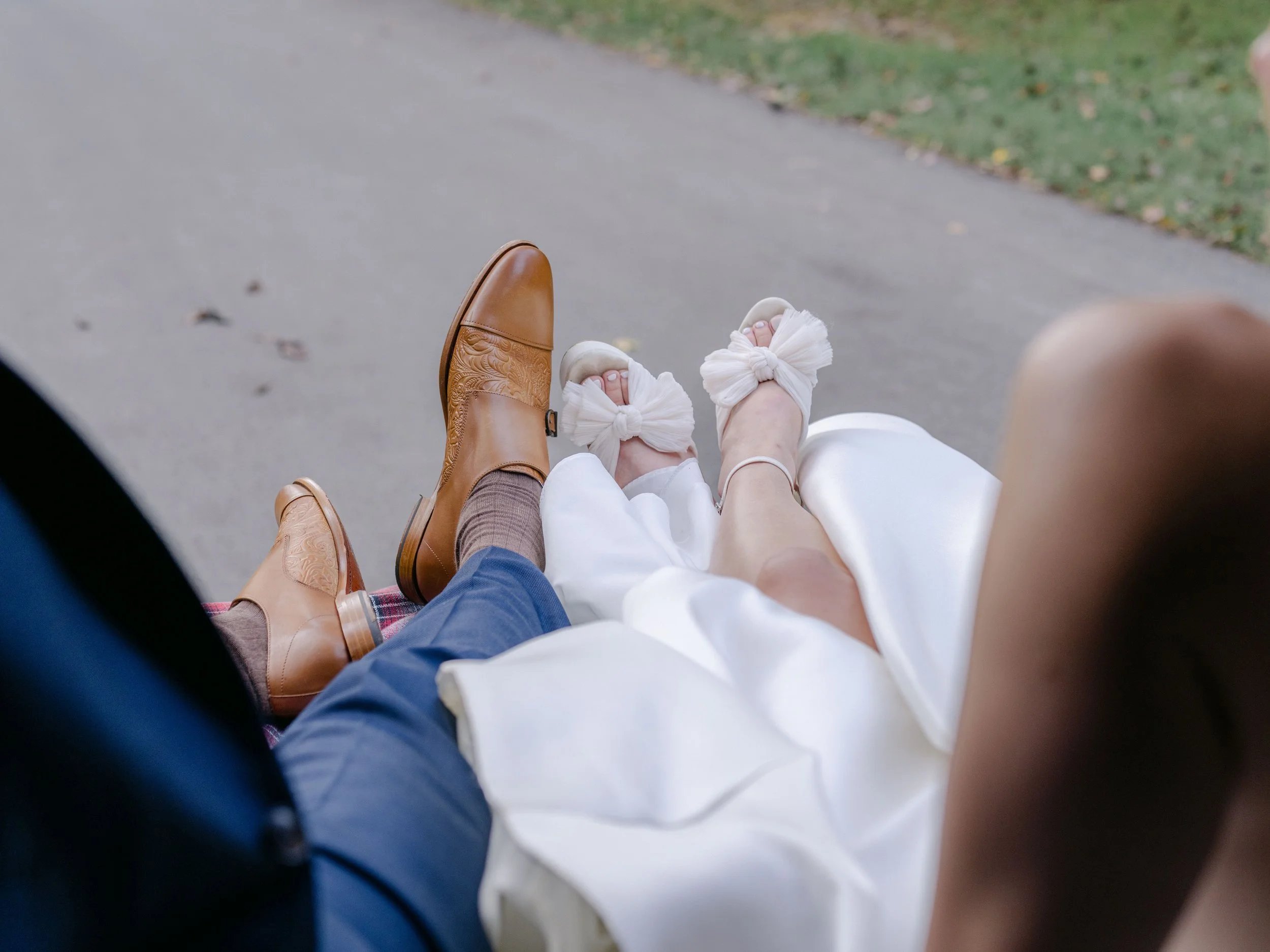 Couple riding in a carriage, showcasing their feet; one person is wearing brown cowboy boots with blue pants, and the other is in white satin shoes with large bows and a white dress.