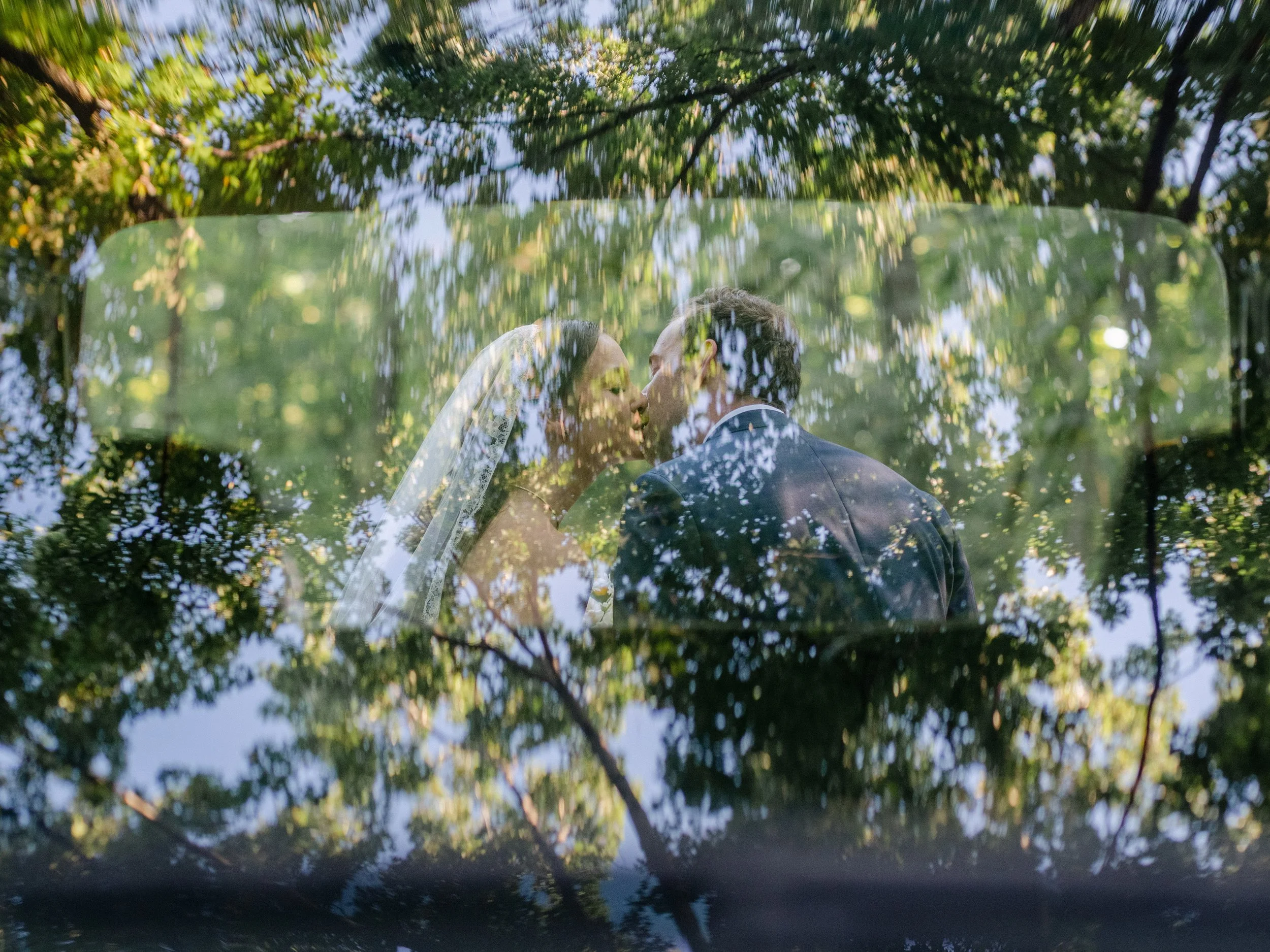 A couple on their wedding day sharing a kiss, with reflections of trees and leaves visible on a glass surface in front of them.