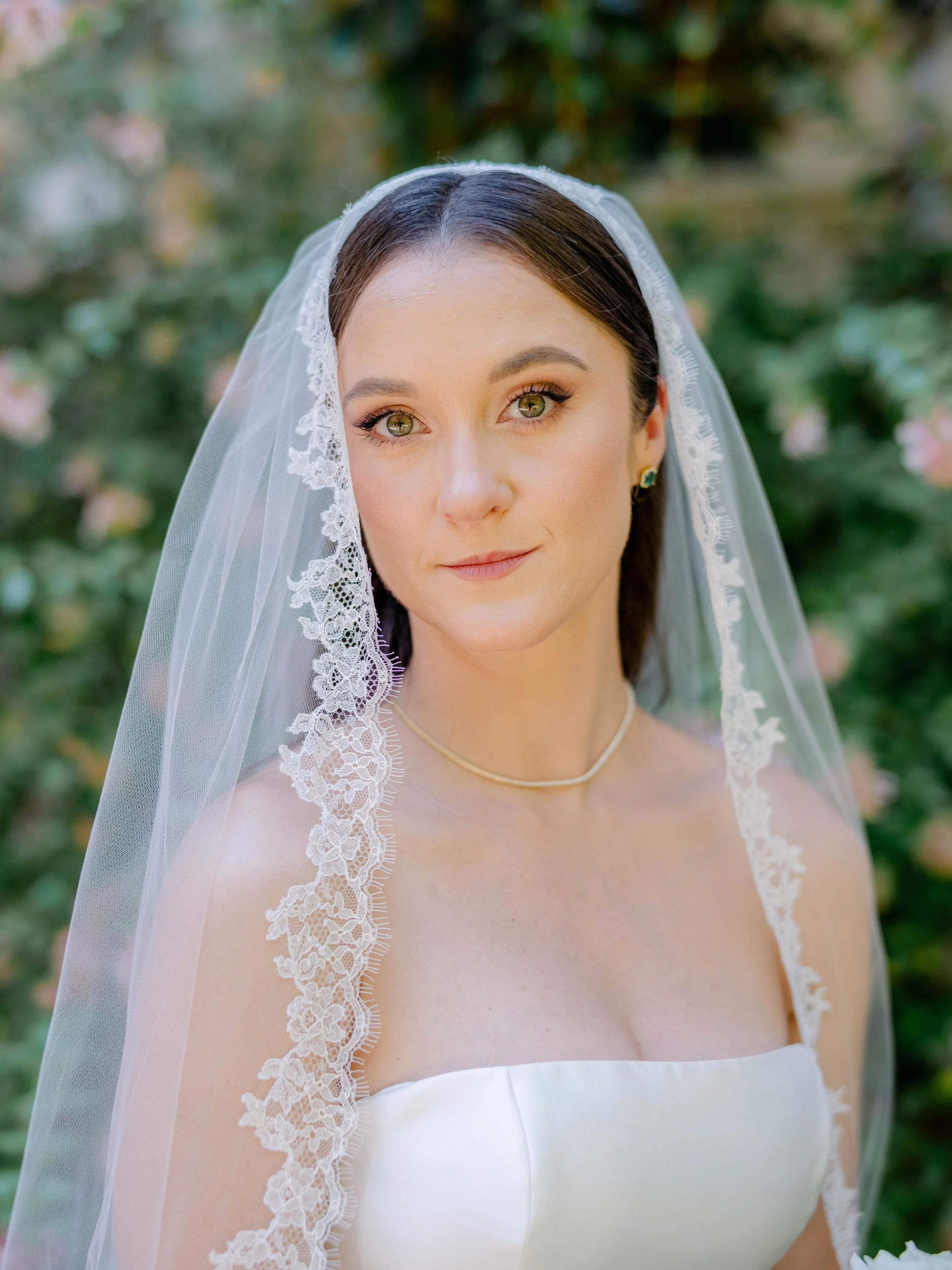 A bride with dark brown hair and green eyes looks directly at the camera. She is wearing a white strapless wedding dress, a delicate necklace, and small earrings. Her sheer veil with lace trim covers her head and shoulders. The background is blurred 