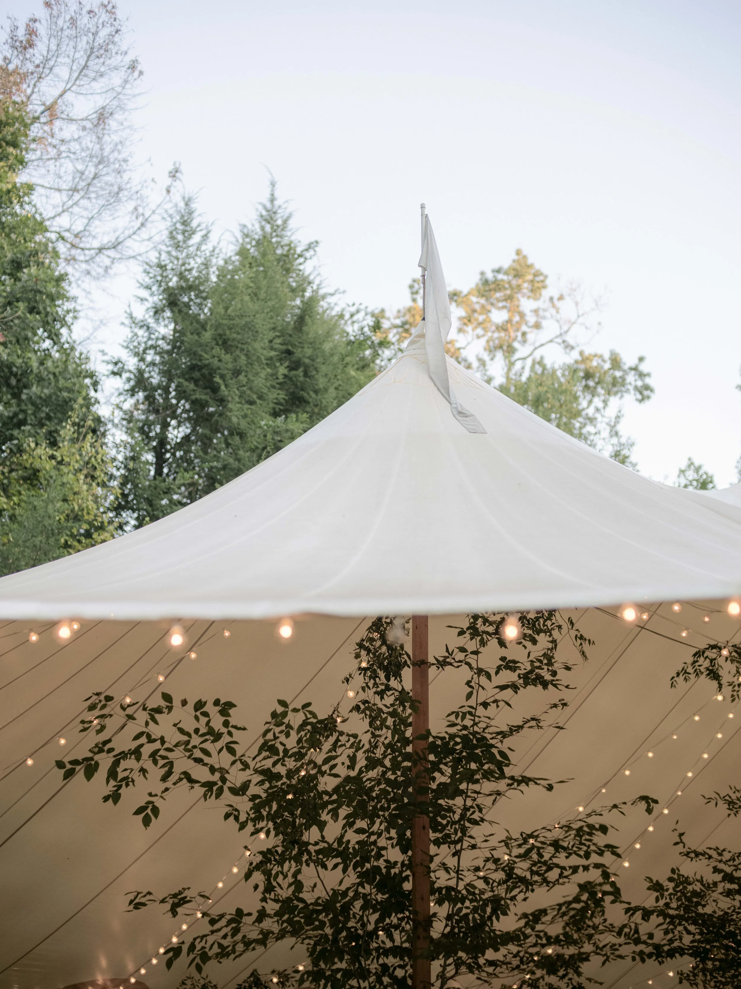 Large beige outdoor event tent with string lights, surrounded by trees, during daytime.