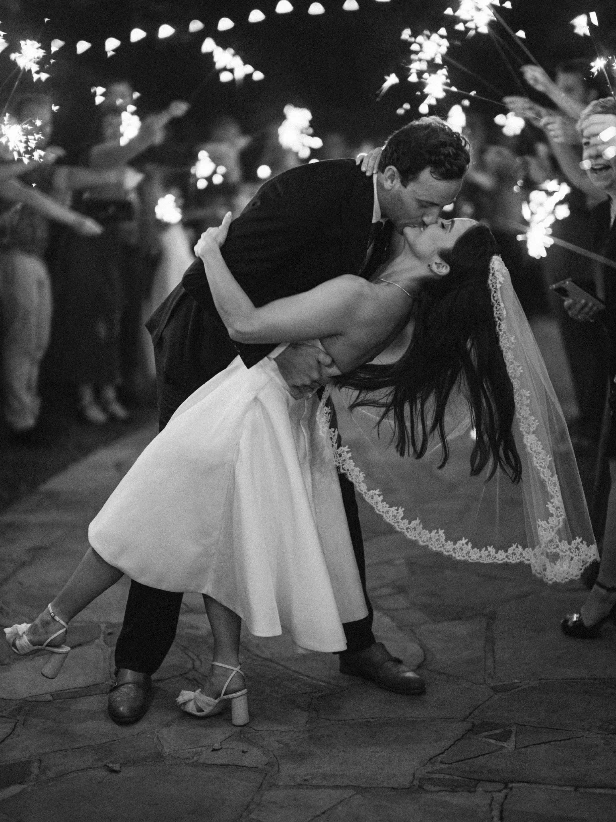 A newlywed couple shares a kiss during their wedding celebration, with guests holding sparklers in the background.