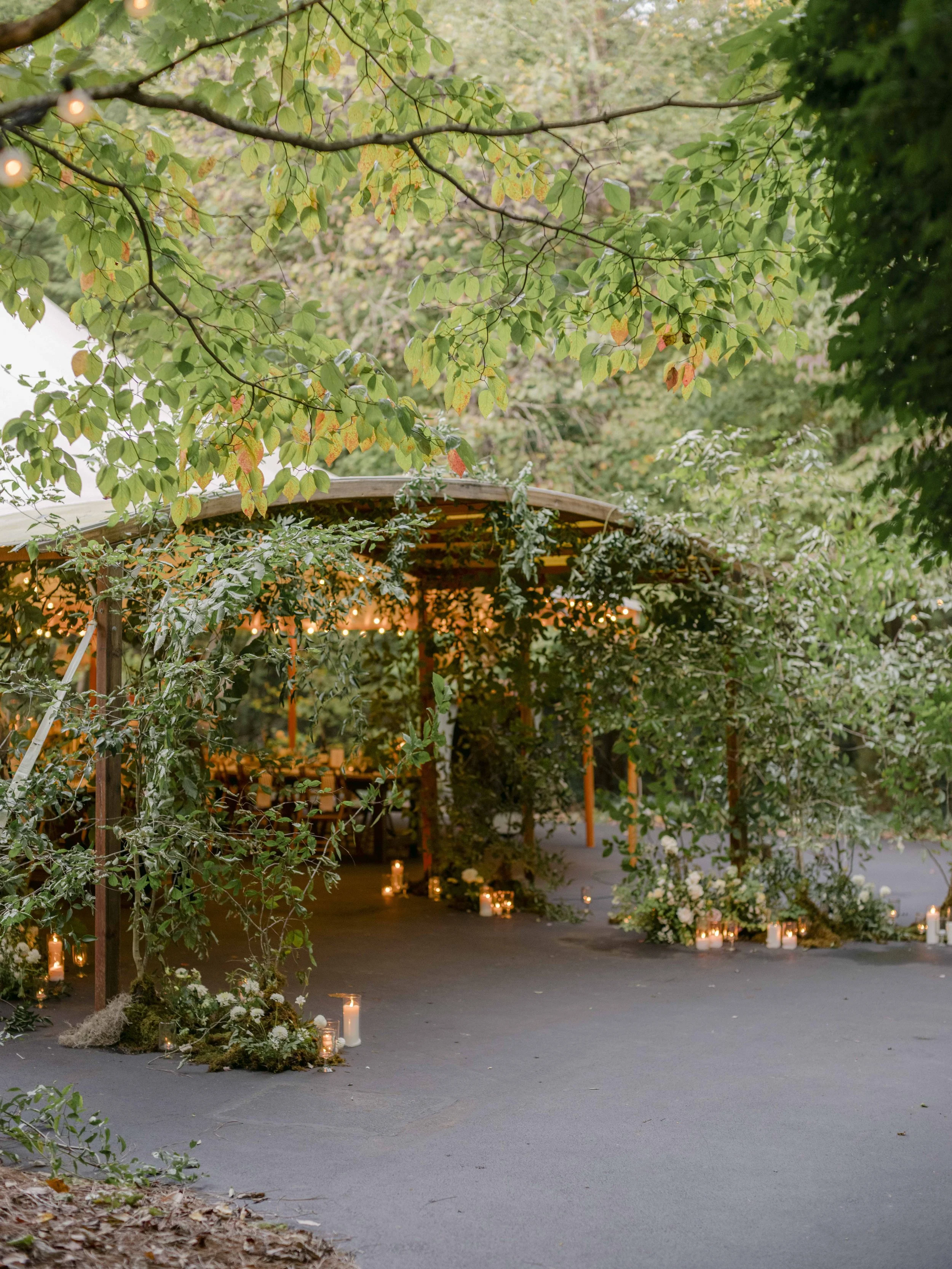 Outdoor wedding reception area decorated with candles and lush green foliage, under a wooden canopy.