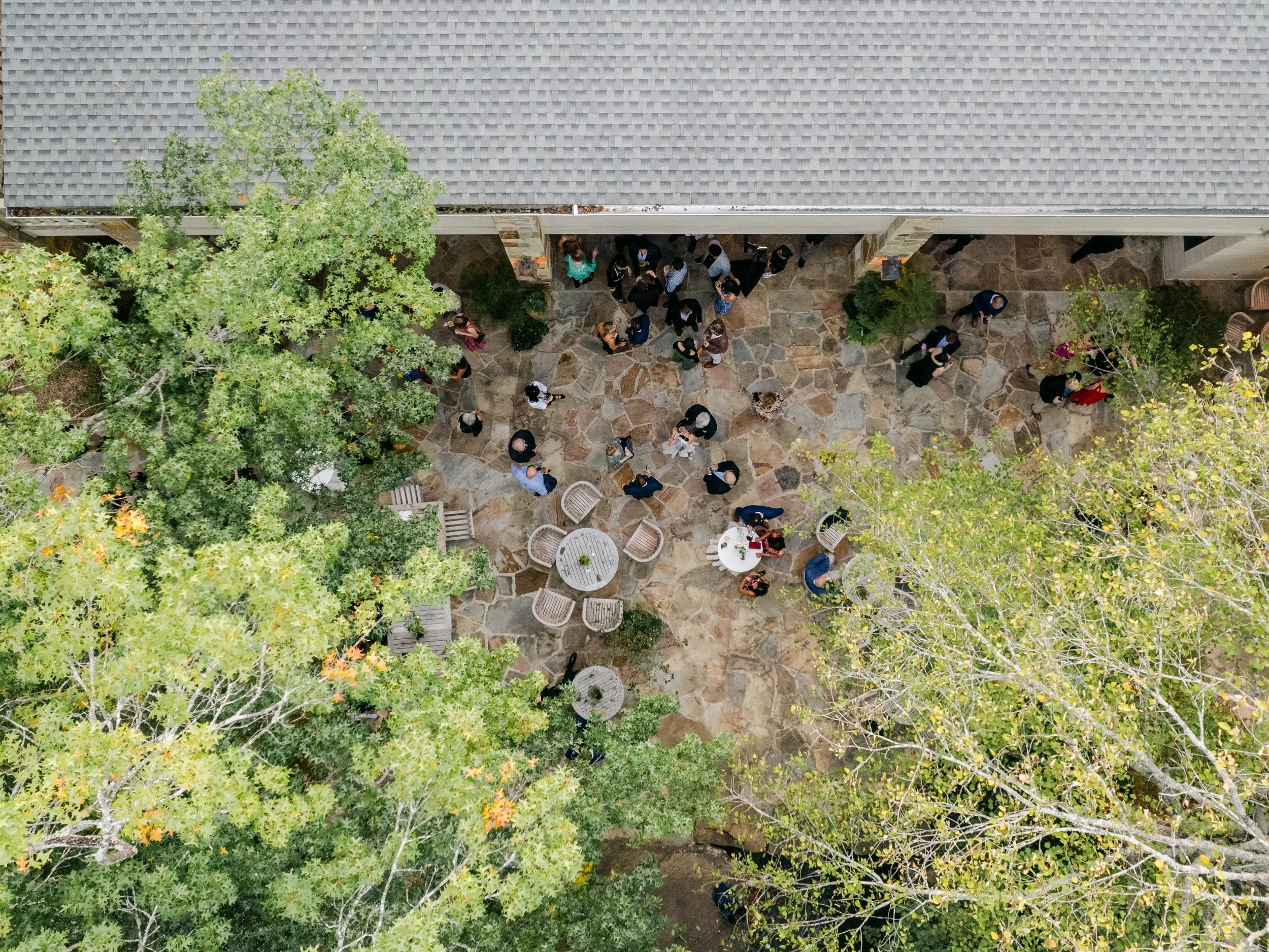 An aerial view of people gathered outside a building, on a stone patio surrounded by trees and outdoor seating.