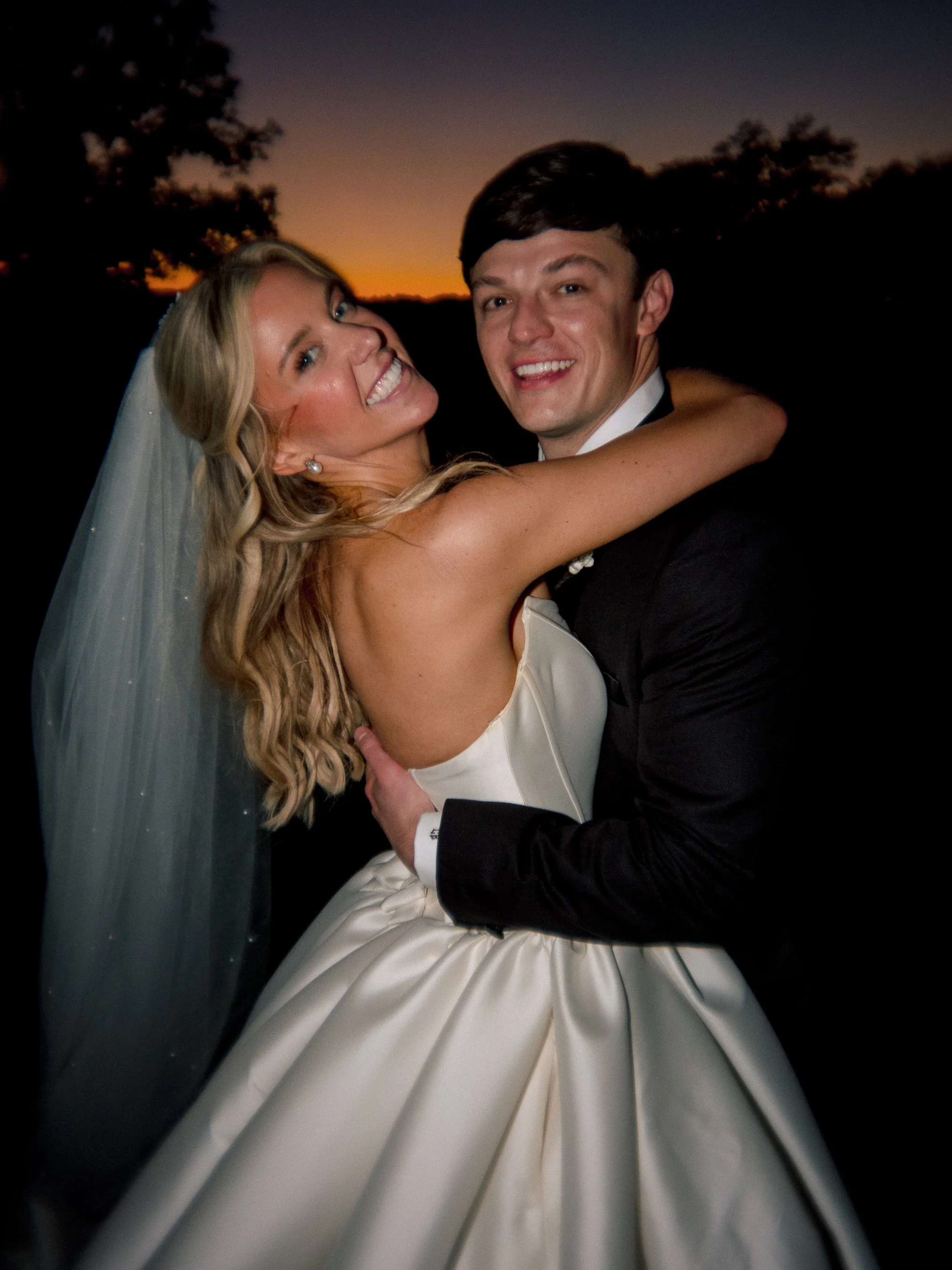 Happy couple, bride and groom, embracing outdoors at sunset, smiling, wedding attire, bride in wedding dress with veil, groom in tuxedo.