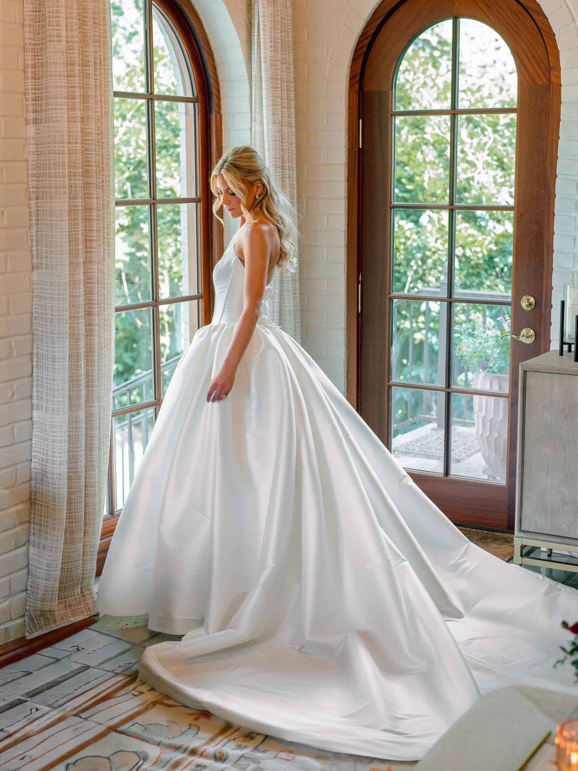 A woman in a white wedding gown standing by a window with natural light streaming in, in a room with white brick walls and wooden framed glass doors.