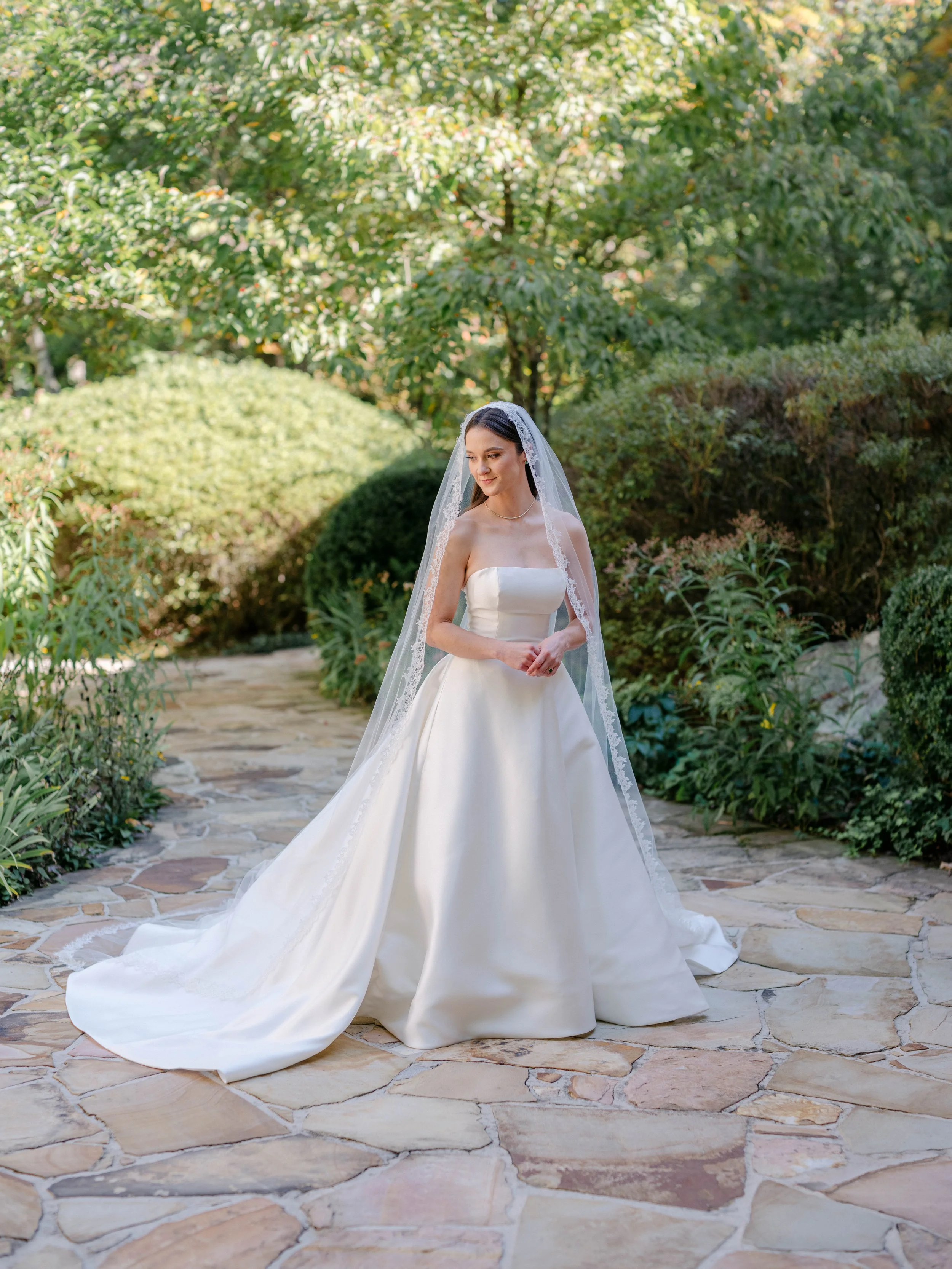 A bride in a white wedding gown with a veil standing on a stone pathway surrounded by greenery in an outdoor garden setting.
