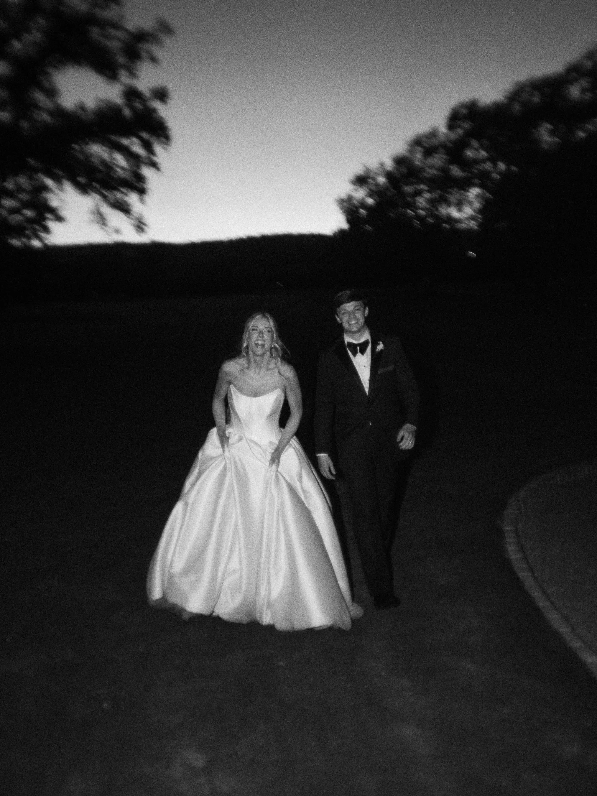 A bride in a strapless wedding gown and a groom in a tuxedo walking outdoors at dusk.