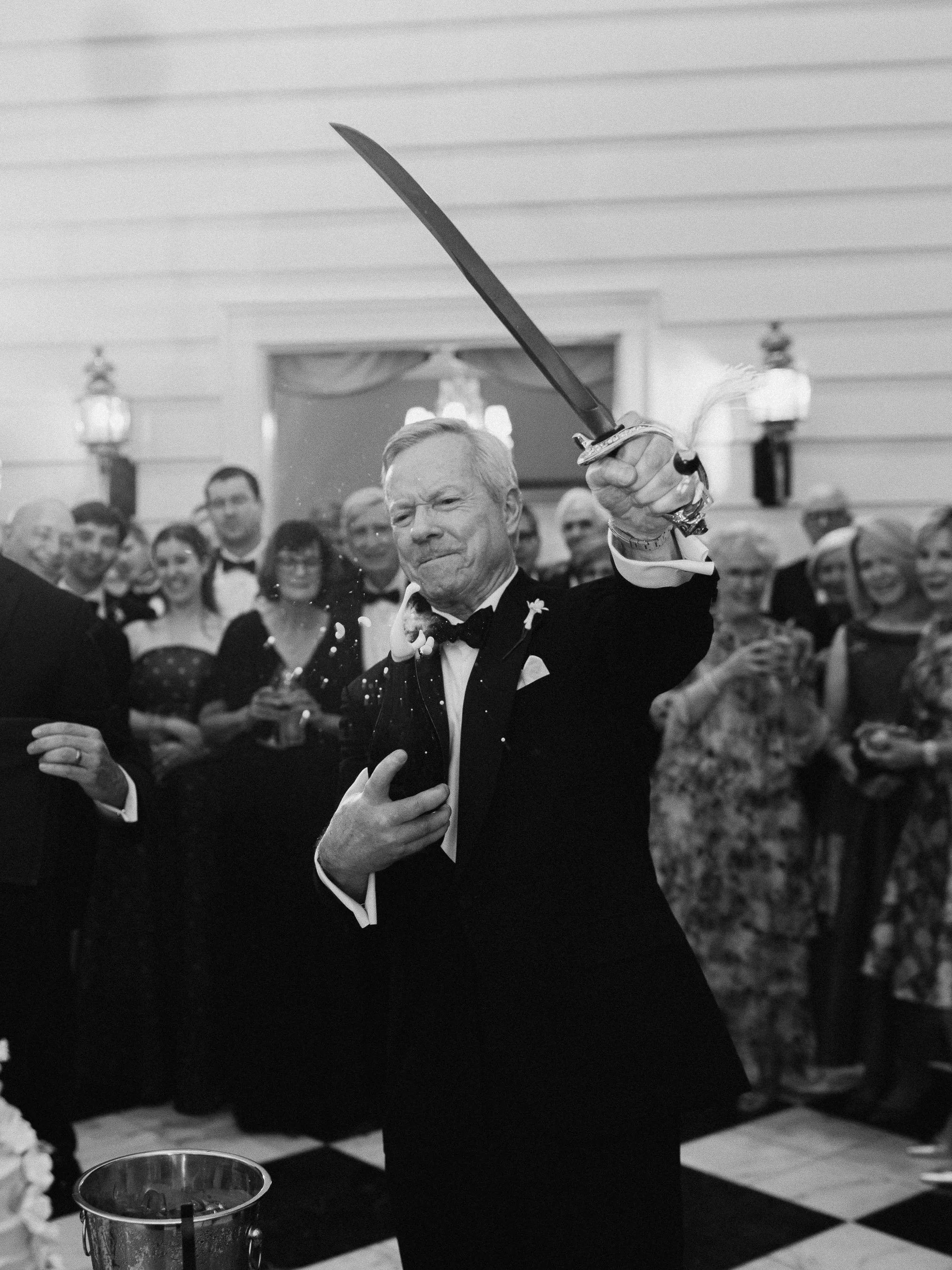 An older man in a tuxedo holding a sword, standing in front of a group of people at a formal event. The scene appears to be a celebratory or ceremonial moment.