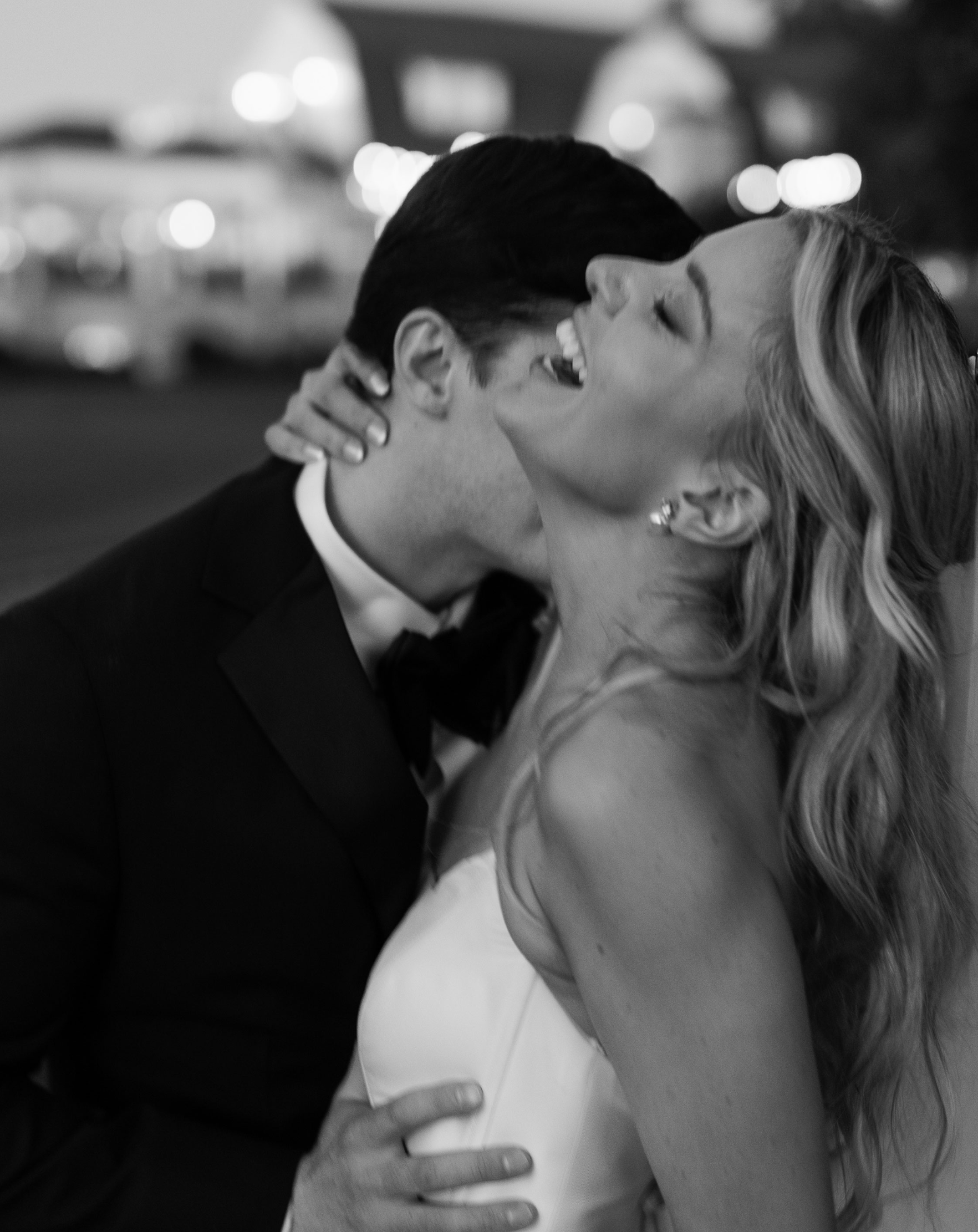 A black and white photograph of a couple sharing a kiss at night. The man is dressed in a tuxedo and the woman is in a white wedding dress with her eyes closed, smiling.