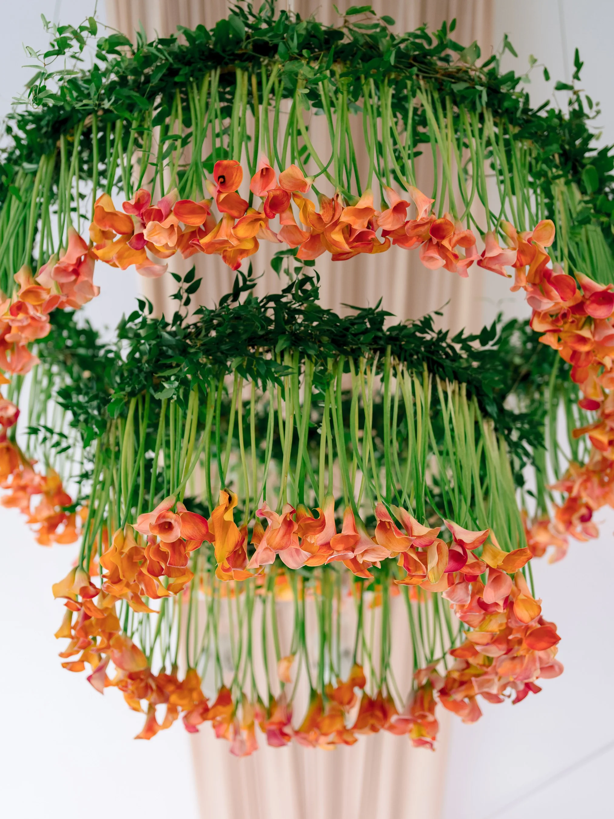 Hanging light fixture made of greenery, orange and pink flowers, and green stems, with a blurred neutral background.