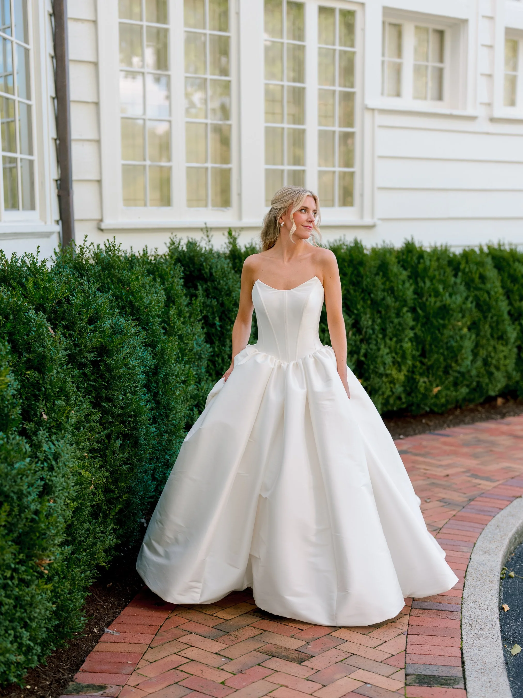 A woman wearing a white strapless wedding gown with a full skirt, standing outdoors on a brick walkway, with green shrubbery and a white building with large windows in the background.