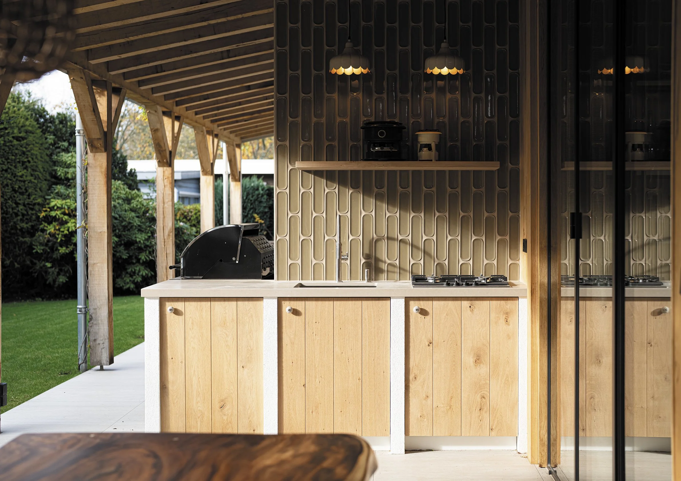 Buitenkeuken met houten kasten, een gootsteen, een gasfornuis en een afzuigkap, met een houten plafond en groene bomen op de achtergrond.