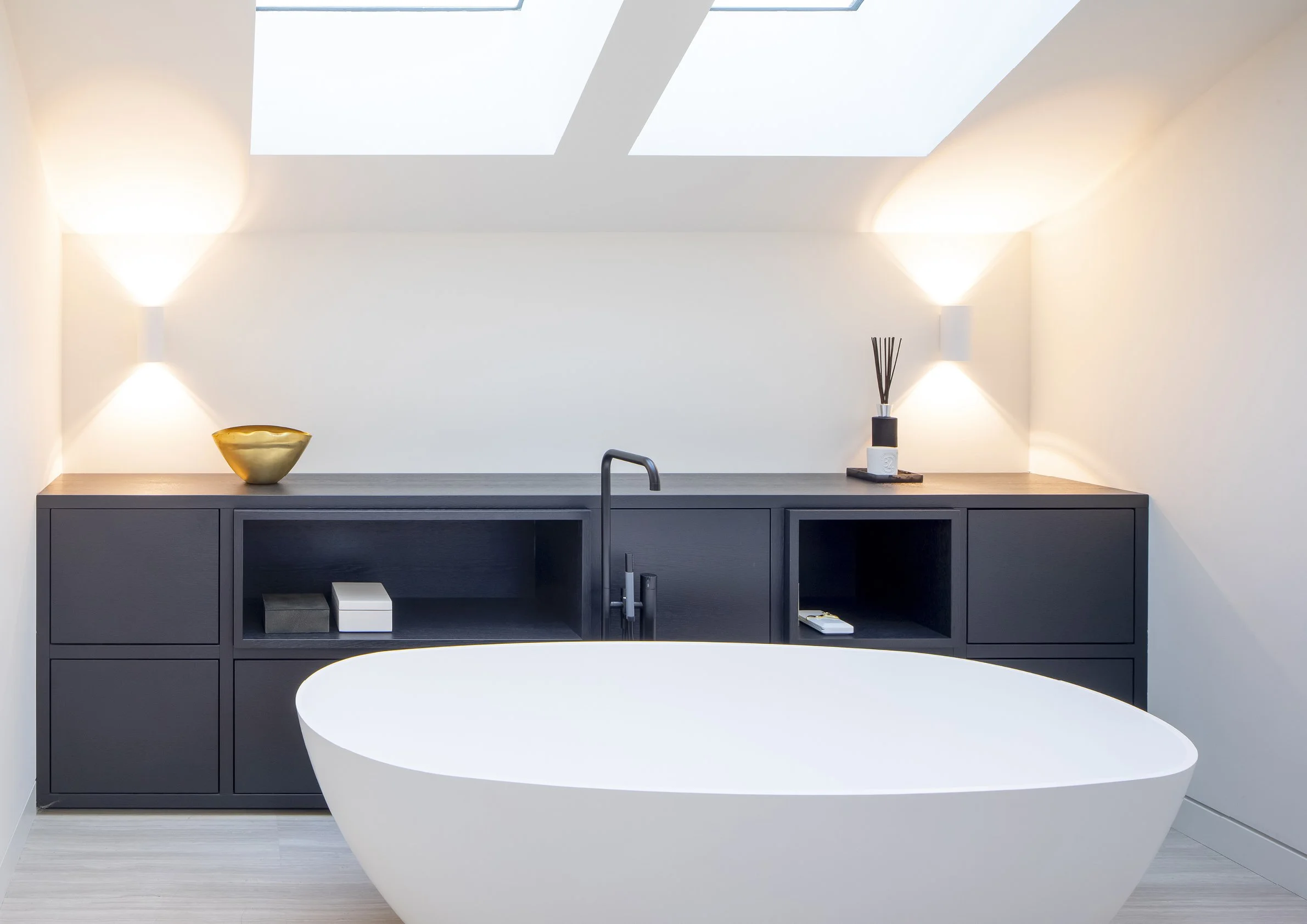 Een witte vrijstaande badkuip in een modern badkamersinterieur met een donkere kast en twee wandlampen