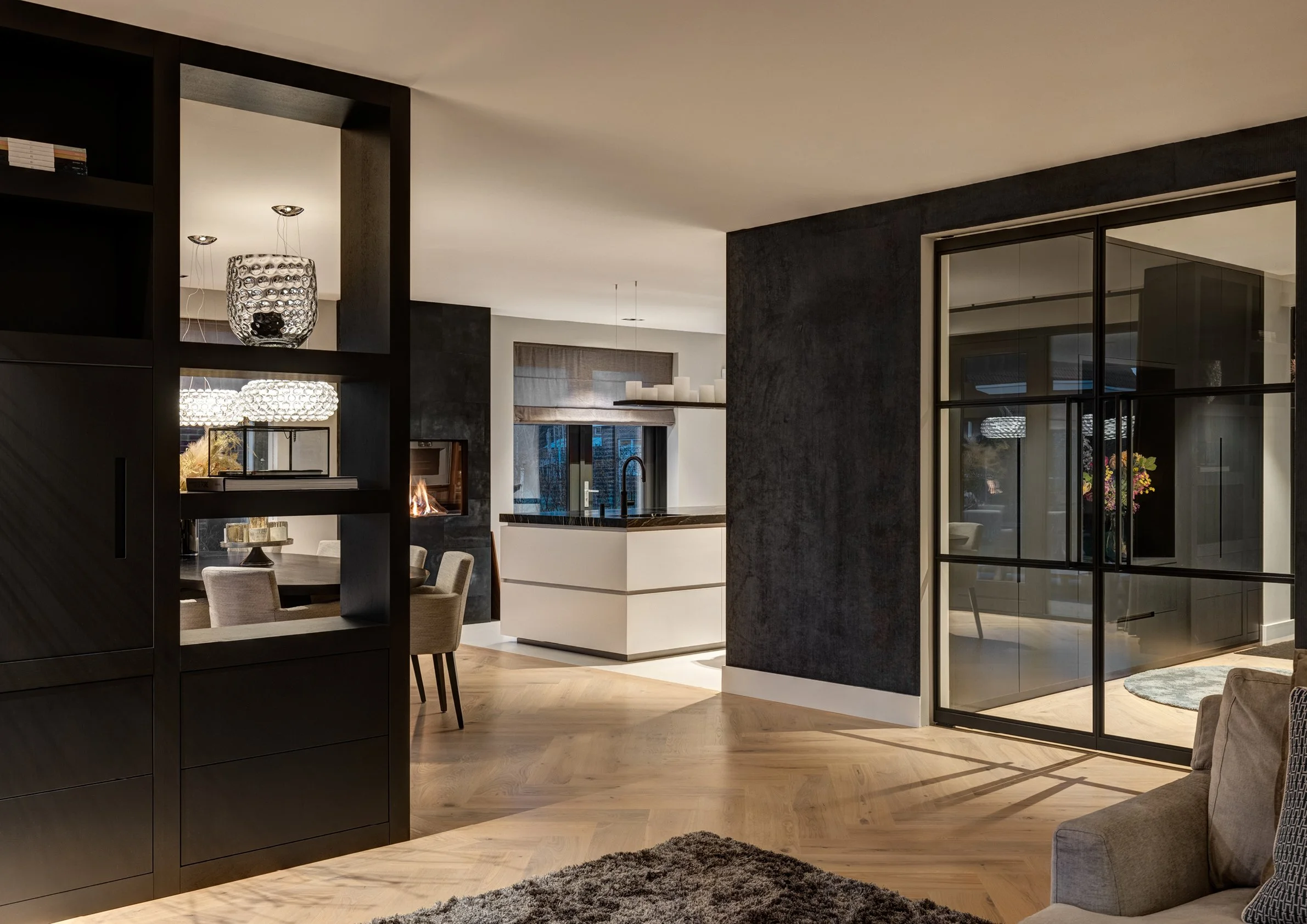 Een modern interieur met een open keuken, eet- en woonkamer, met donkere en lichte elementen, houten vloer en grote ramen.