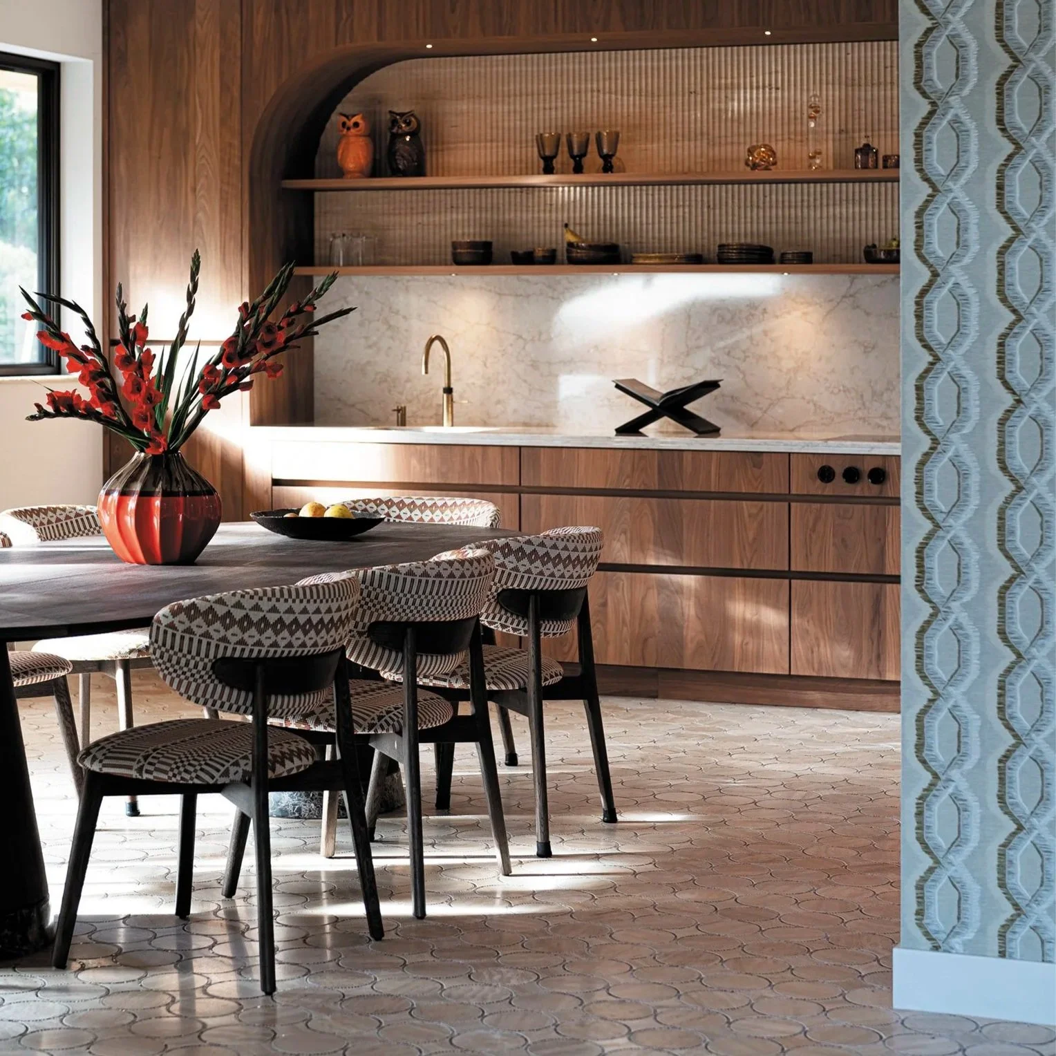 Een moderne keuken en eethoek met een houten tafel, zes eetkamerstoelen met geometrisch patroon, een grote vaas met rode bloemen, openbare houten planken met decoratieve voorwerpen, een witte marmeren keukenachterwand met een gouden kraan, en een jurk met een boek in de keuken.