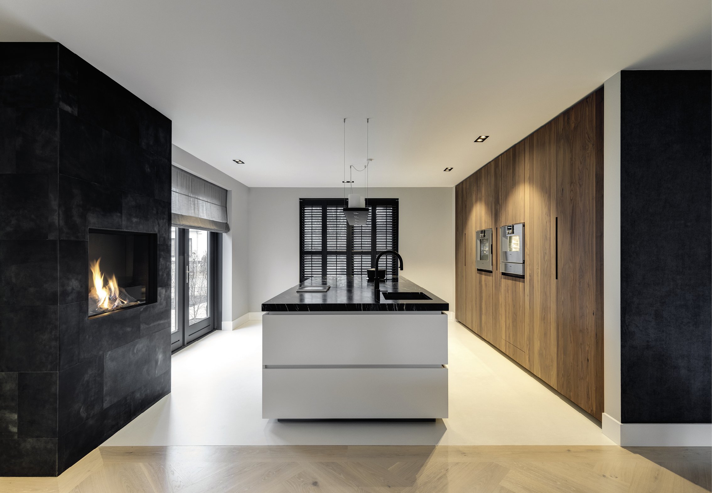 Moderne keuken met zwart aanrecht, houten muur met ingebouwde ovens, zwart raam, open haard op zwarte wand, lichte vloer, grote ramen met vitrage.