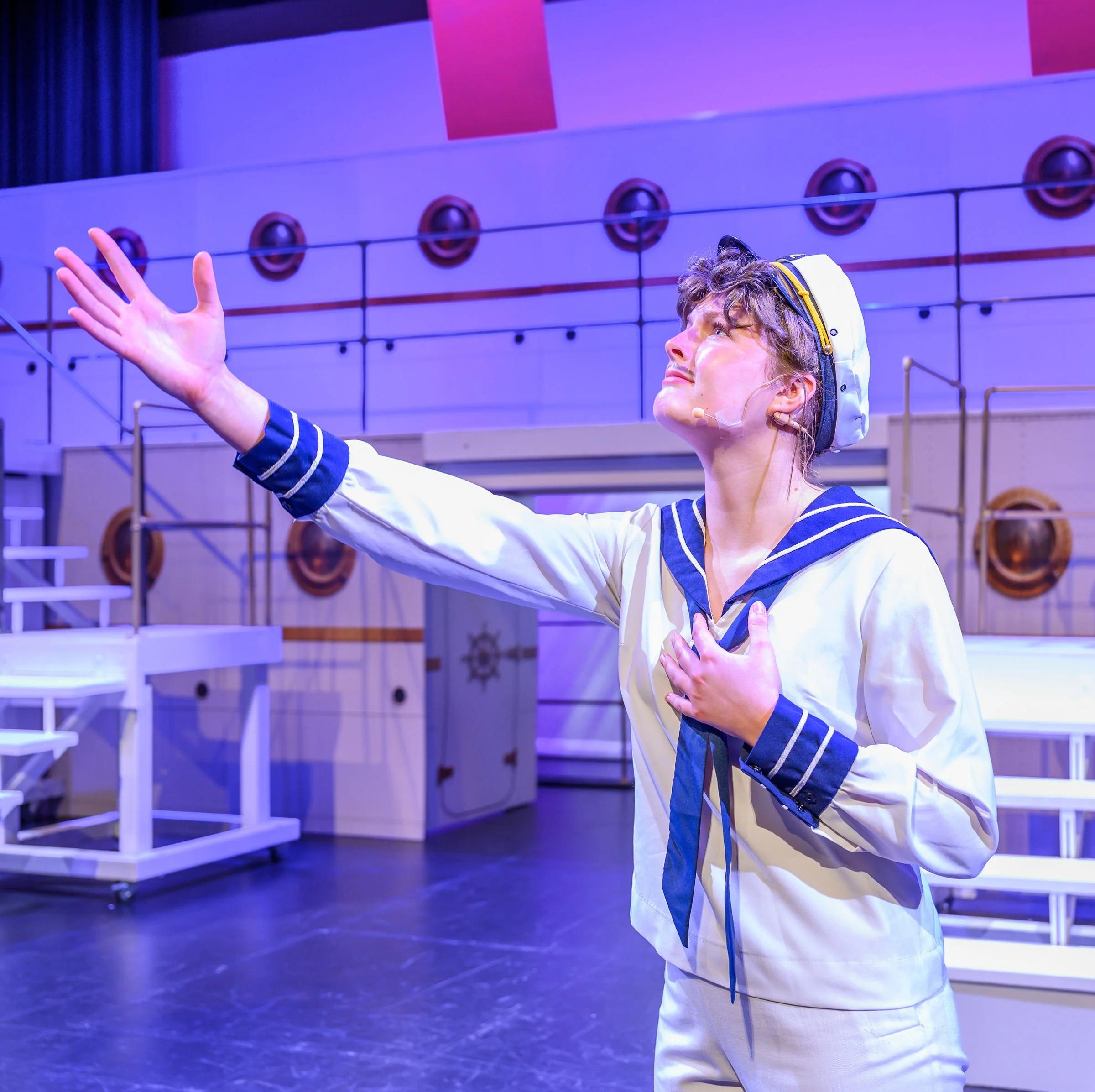 A person dressed as a sailor with a headset and microphone, reaching out with one hand, standing in a room with purple lighting and round objects on the wall in the background.
