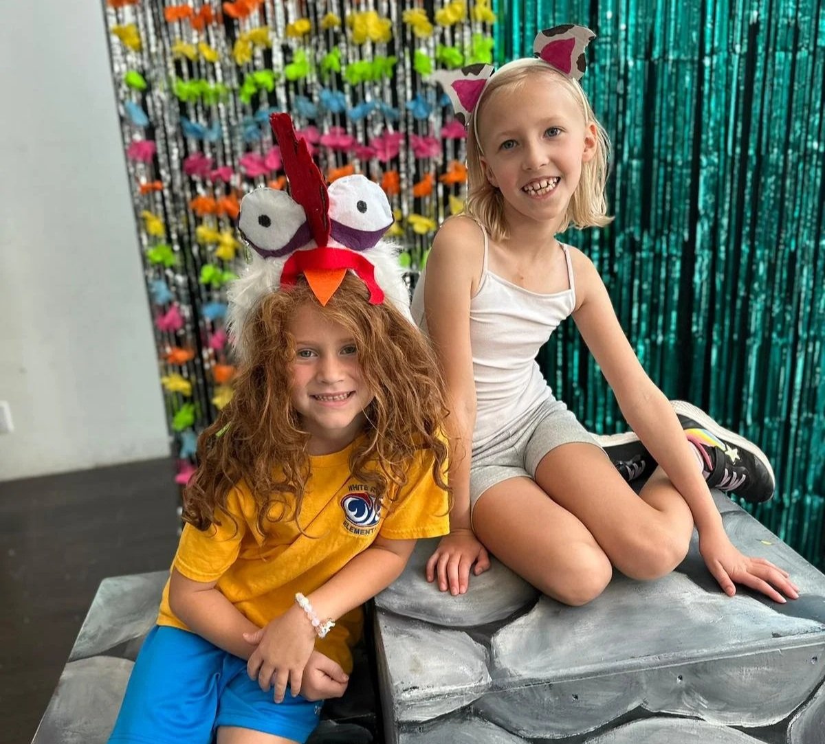 Two young girls sitting on a gray sculpture in front of a colorful butterfly decoration backdrop. The girl on the left has curly red hair, a yellow shirt with a logo, and a turkey headband. The girl on the right has blond hair, a white tank top, and a fox headband.