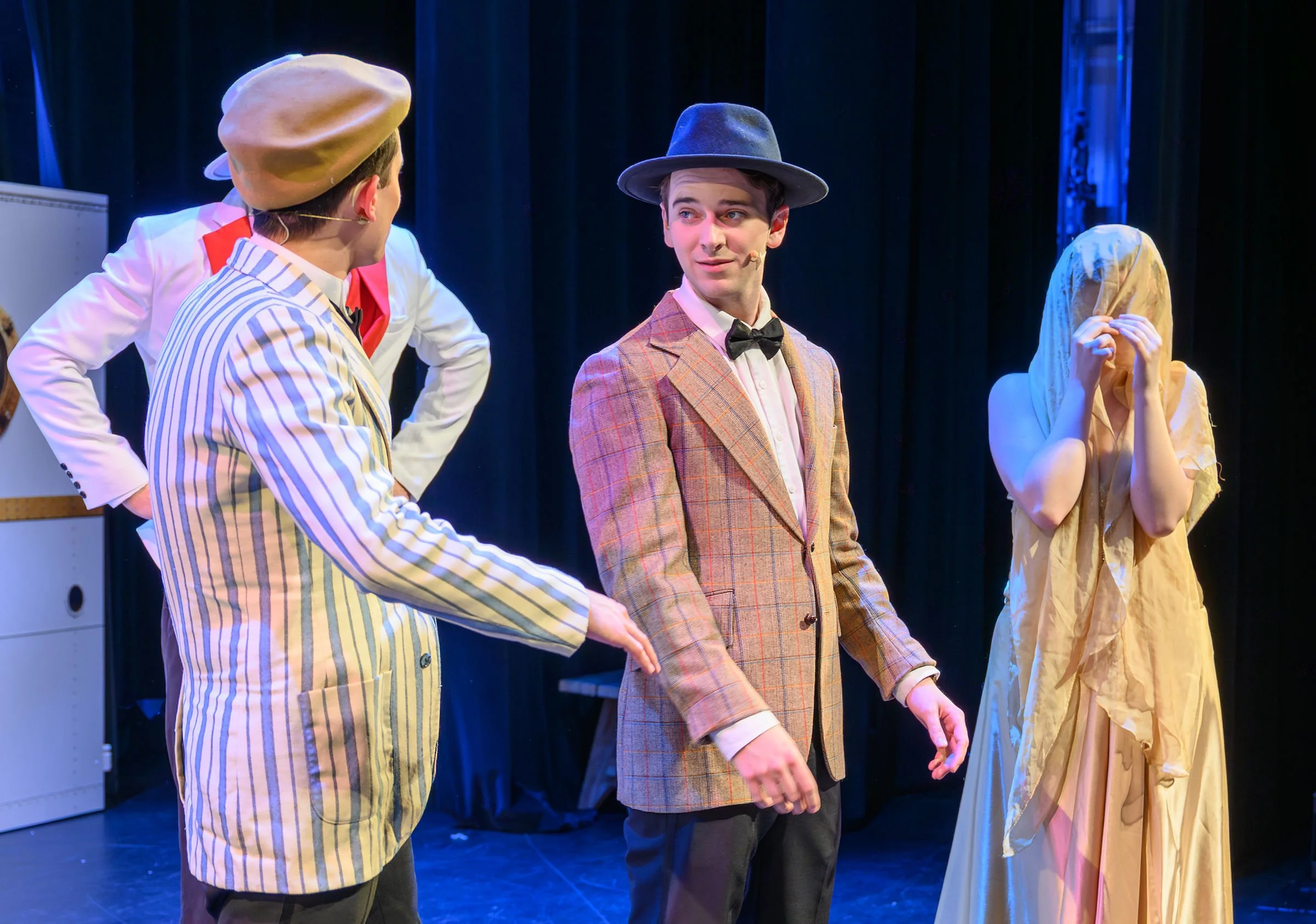 Four actors on stage dressed in vintage costumes, two men wearing suits with bow ties and hats, and two women in dresses with one covering her face with her hands.
