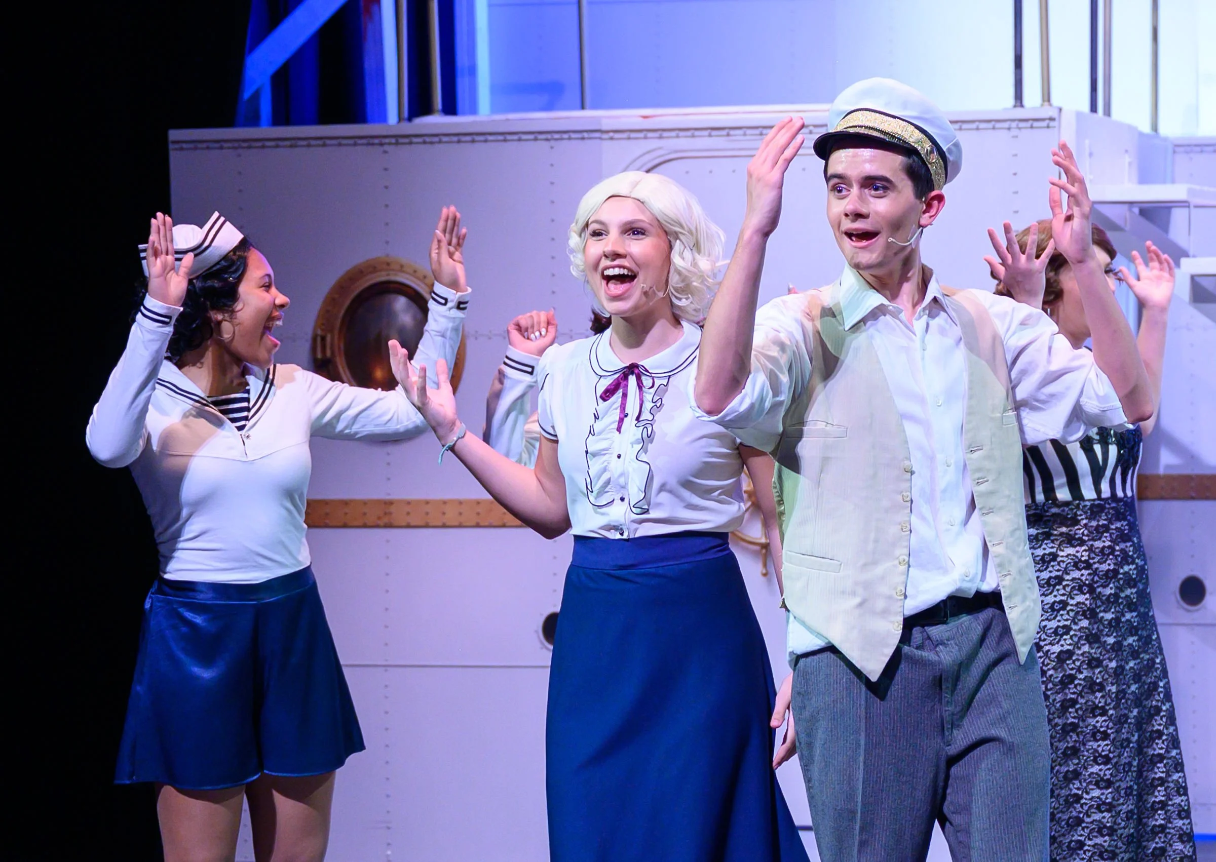Group of young actors dressed as sailors performing on stage, engaging with each other and smiling.