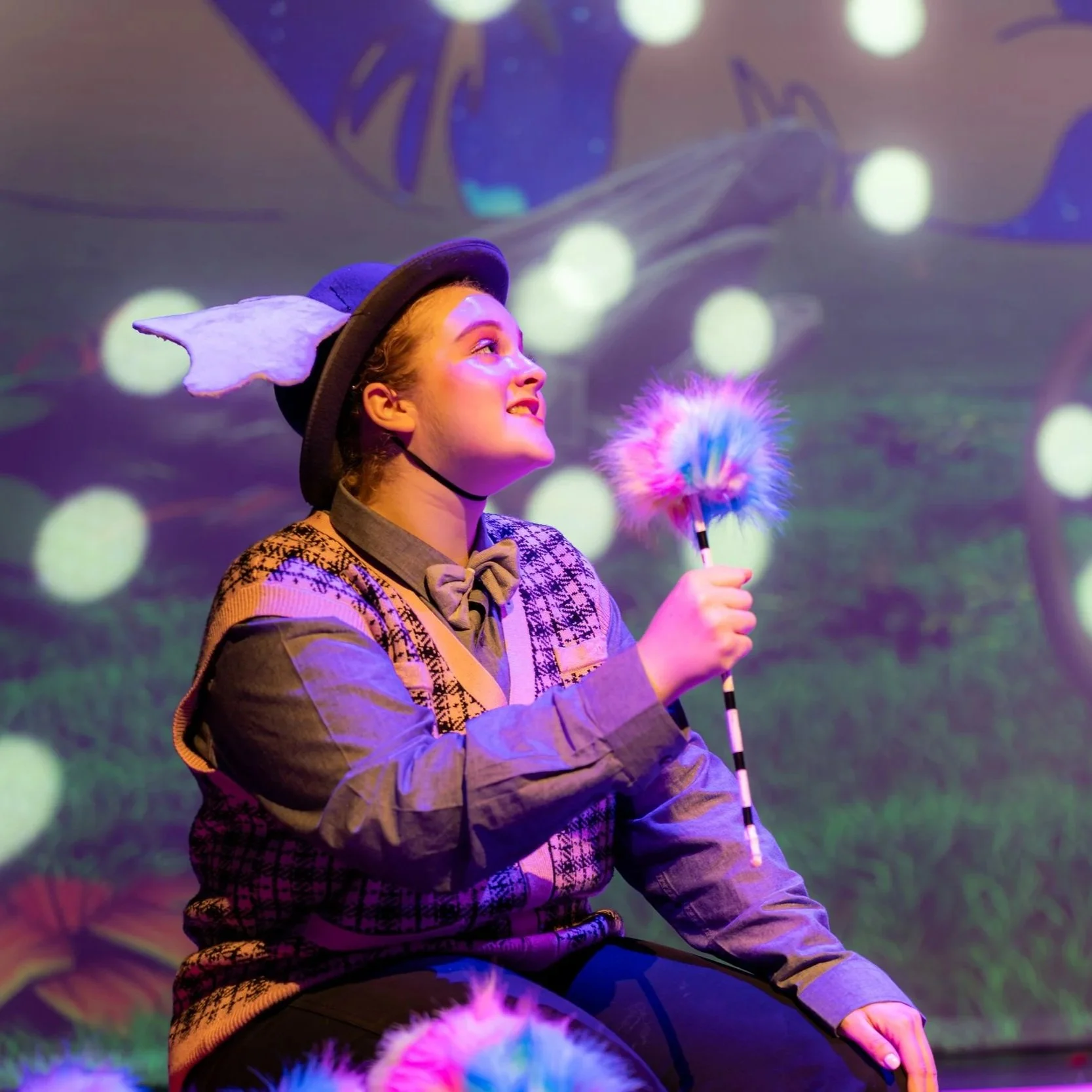 A young woman performing in a colorful theatrical production wearing a costume with a hat and holding a fluffy stick, with a vibrant and whimsical stage background featuring large glowing dots and abstract shapes.