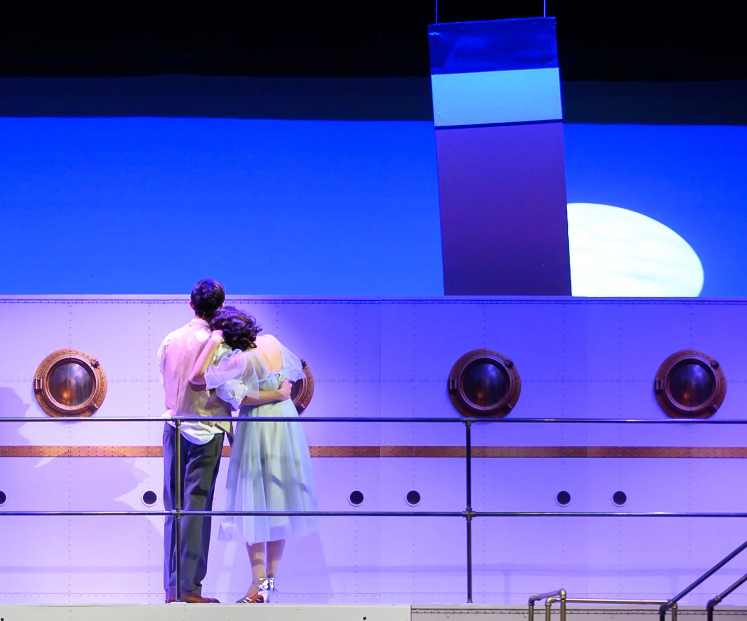 A stage scene with a man and woman holding each other close, dressed in white and light-colored clothing. The background is blue with circular portholes on a white ship or submarine-like structure.