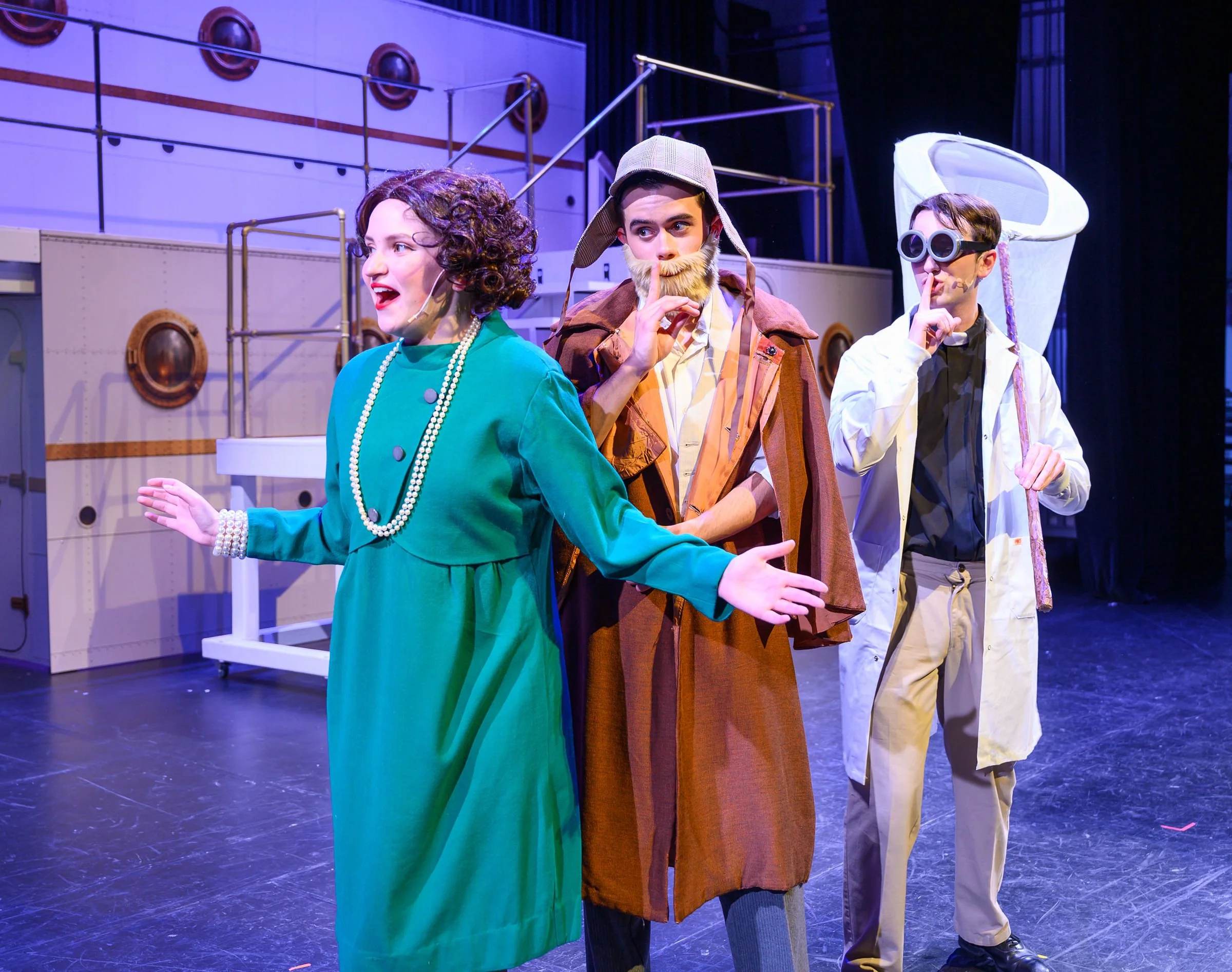 Three actors performing on stage in costumes, with props and set pieces in the background.