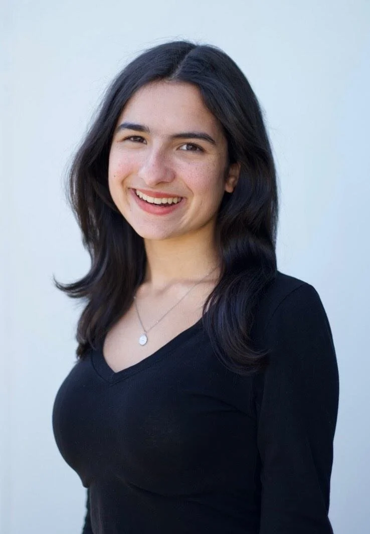 Portrait of a young woman with long dark hair, smiling, wearing a black top and a necklace, standing against a light background.
