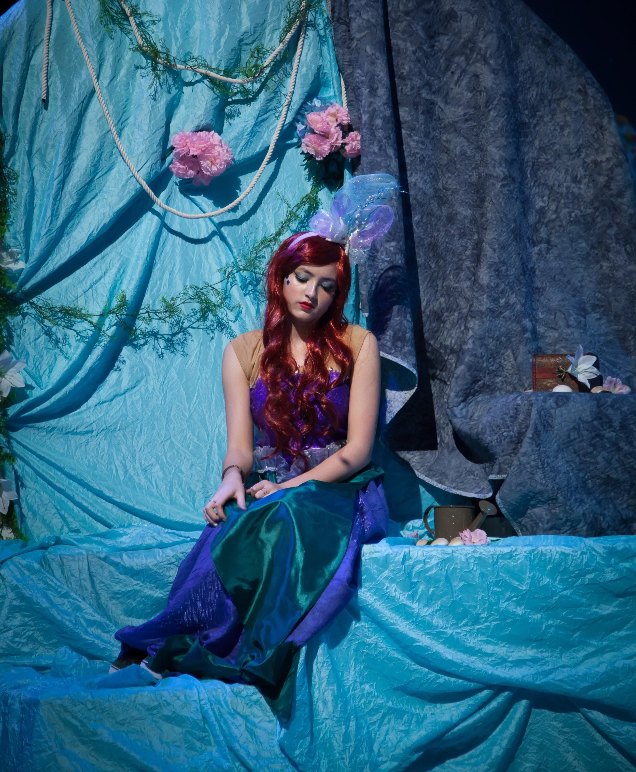 Woman with long red hair sitting on a blue fabric-covered surface surrounded by flowers, draped fabrics, and decorative items, in a fantastical setting.